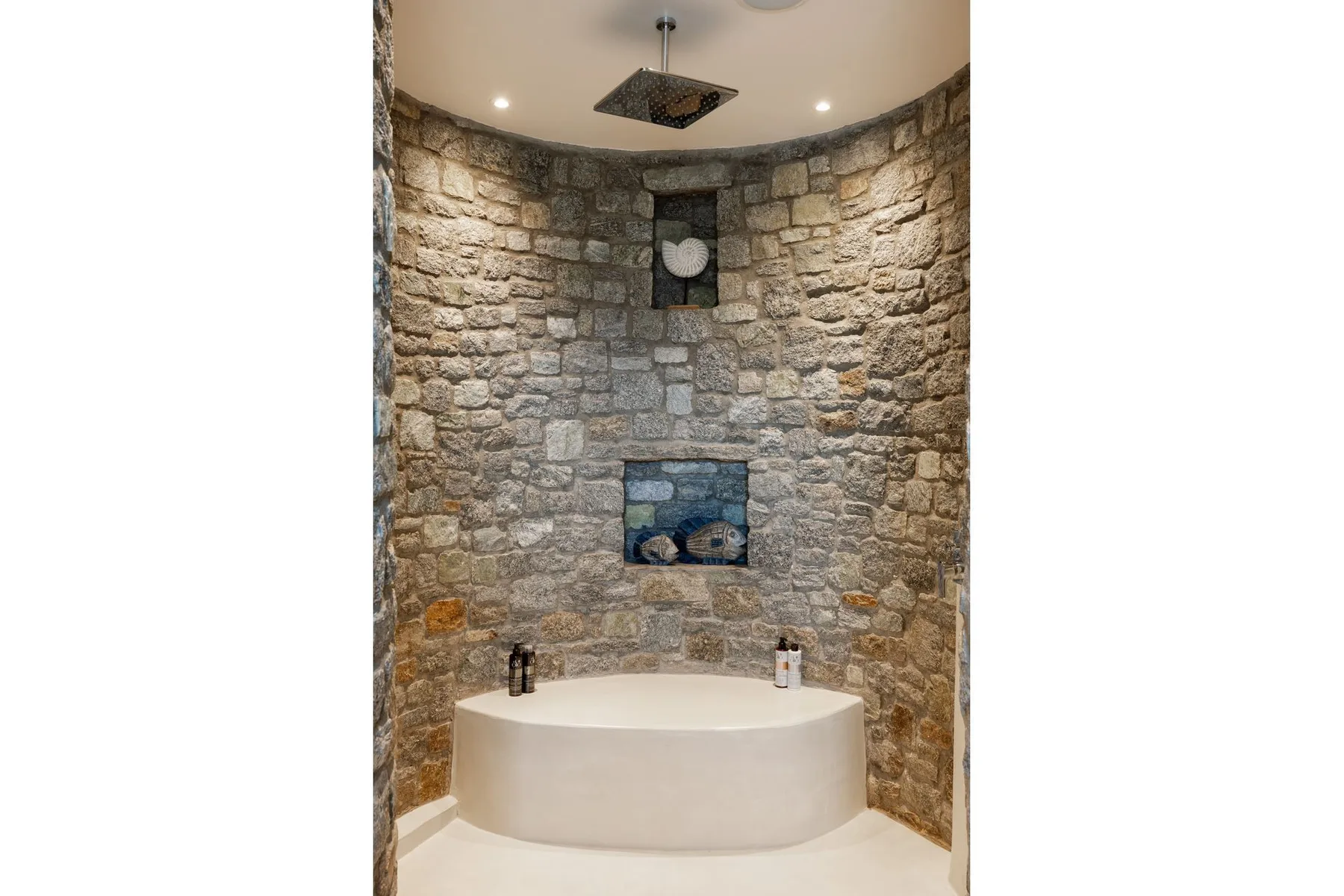 A curved soaking tub sits in a stone-walled bathroom beneath a ceiling showerhead.