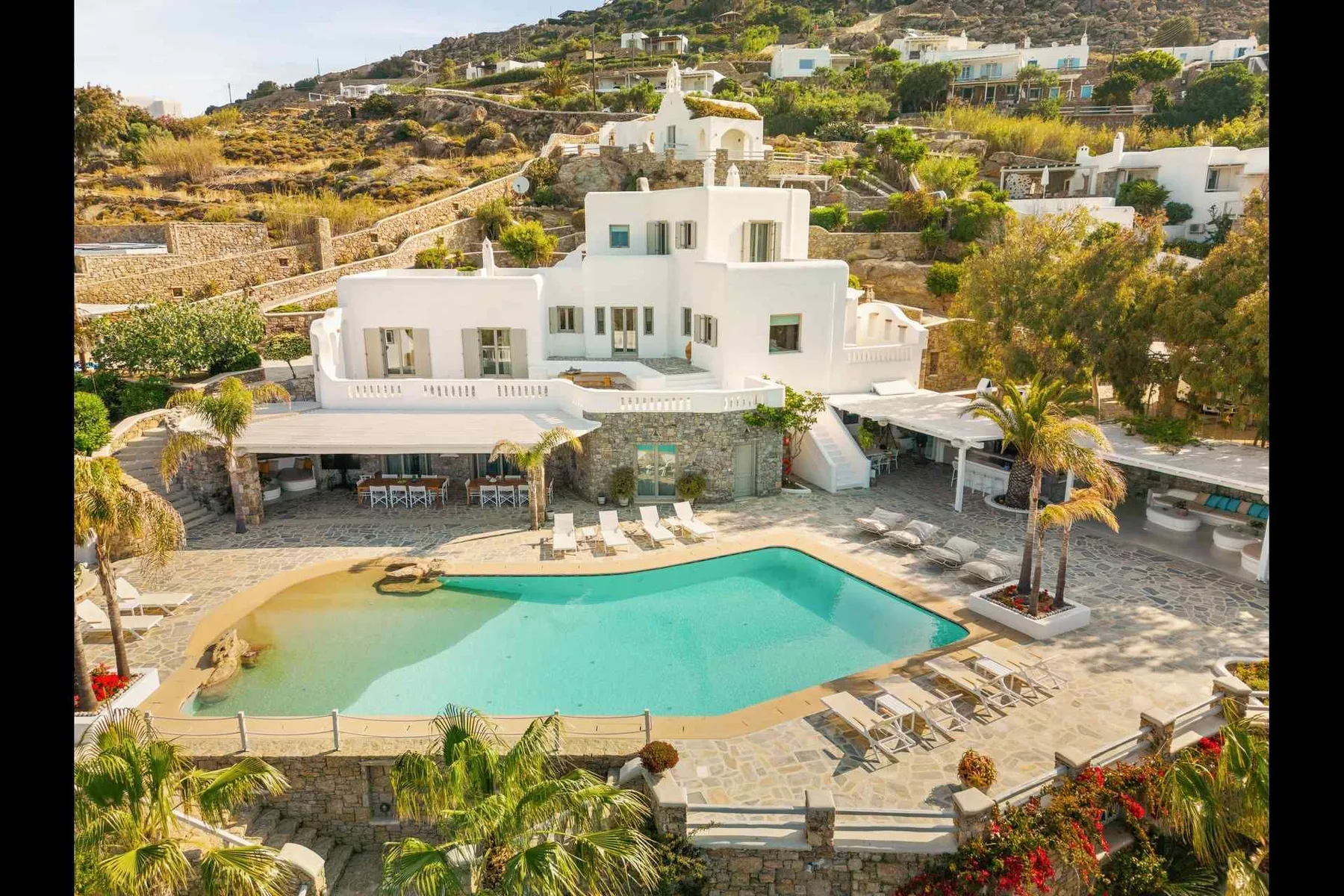 A white hillside villa overlooks a turquoise swimming pool and surrounding terrace.