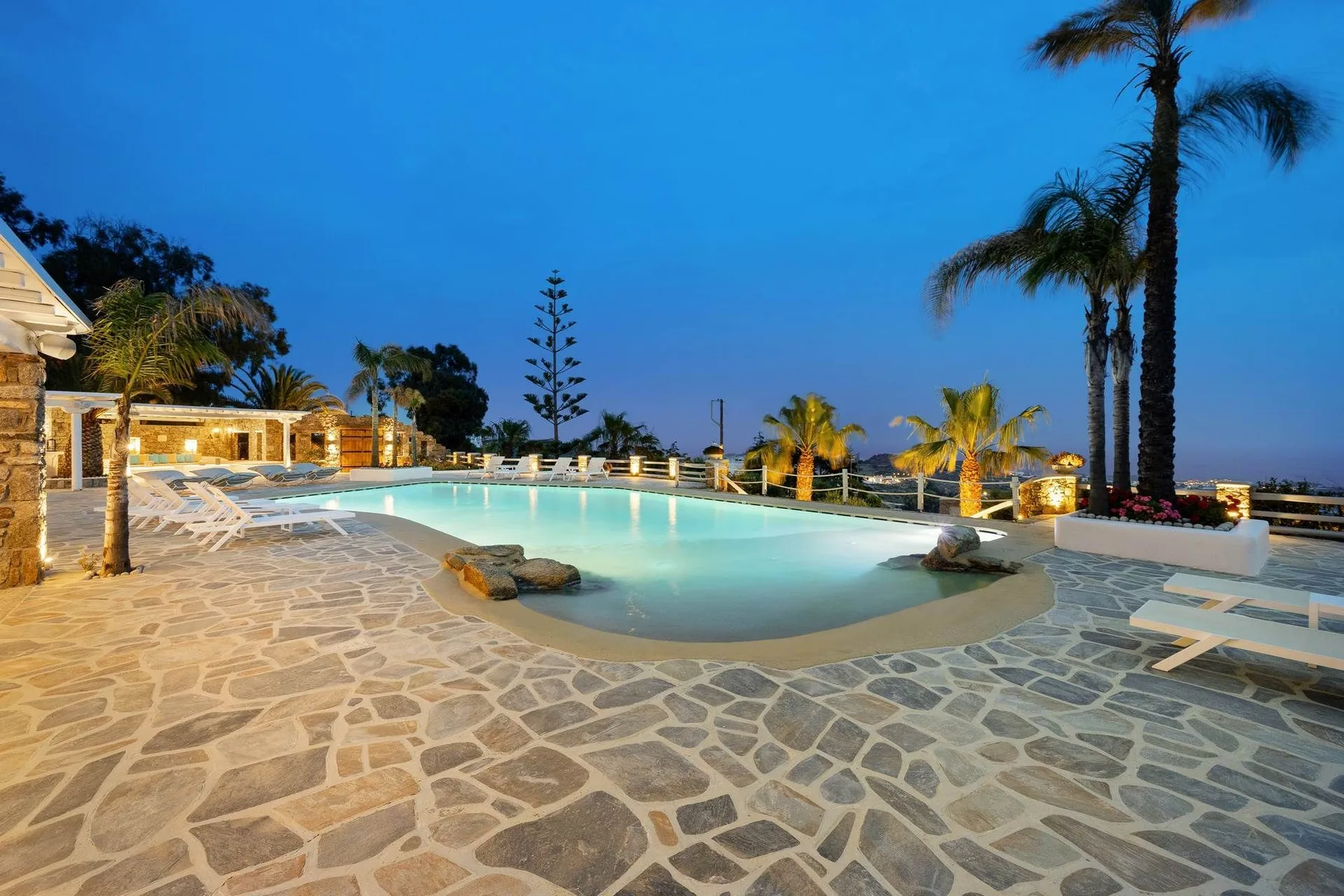 An outdoor swimming pool sits beside a stone-paved terrace at dusk.