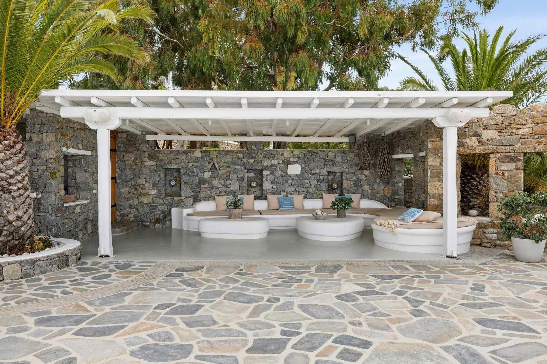 An outdoor lounge beneath a white pergola, with curved seating against stone walls.