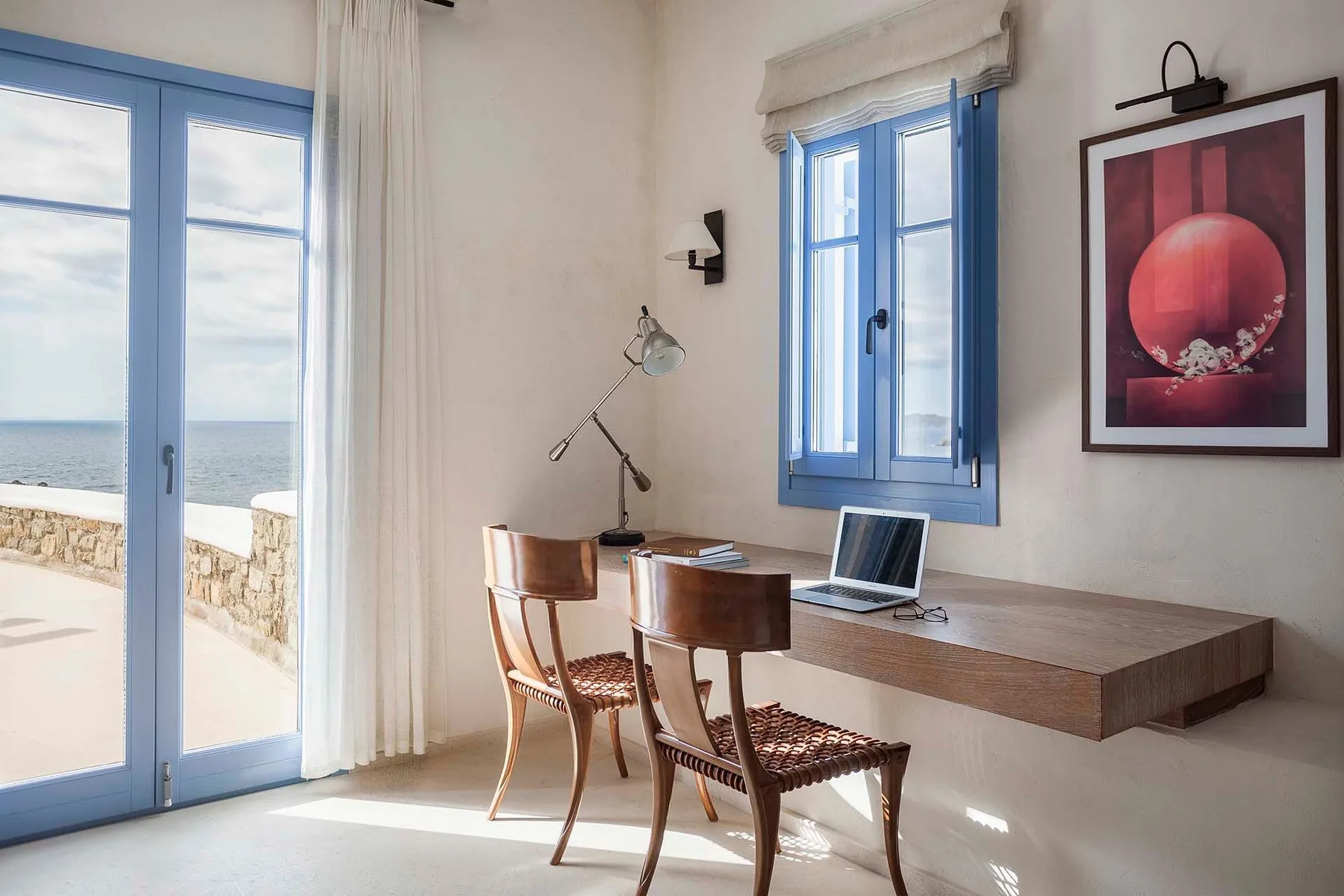 A bright room with a wooden desk and blue-framed windows overlooking the sea.