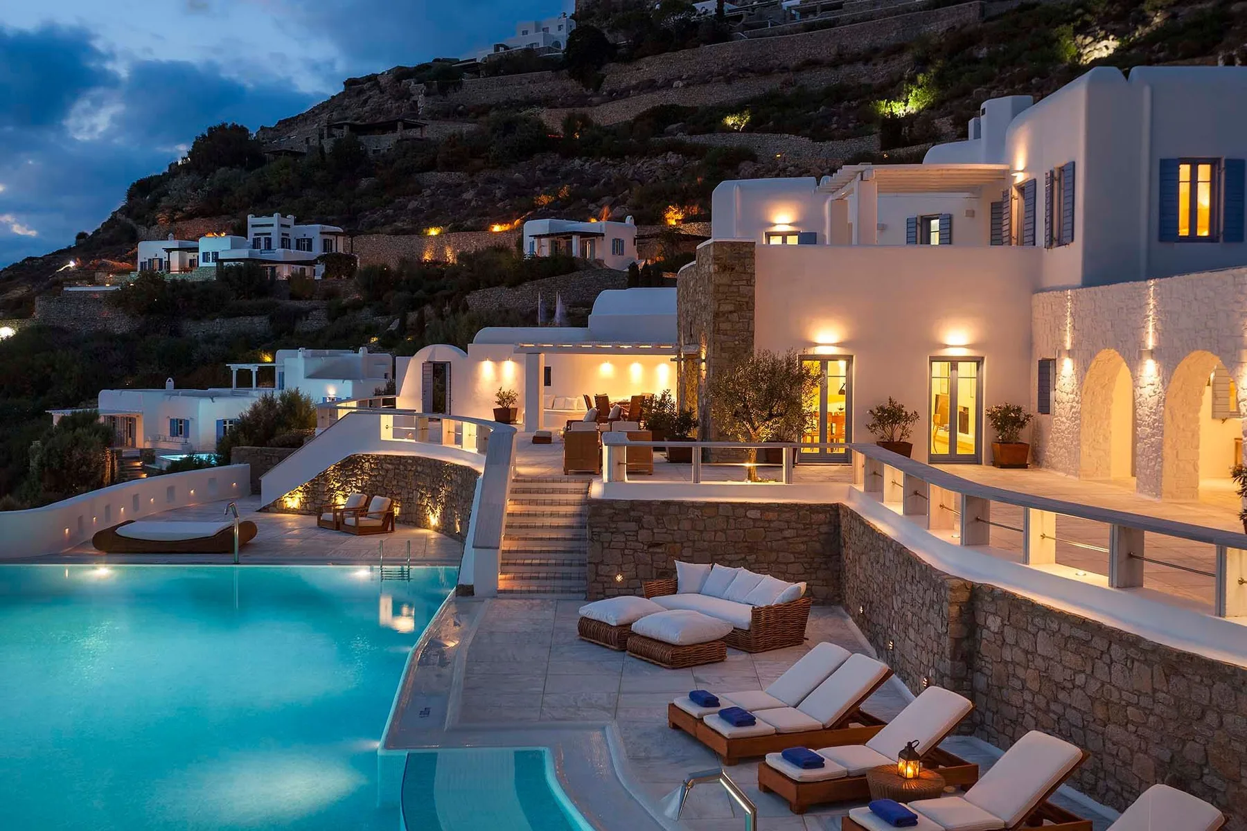 A hillside resort at dusk overlooks a turquoise pool and outdoor seating.