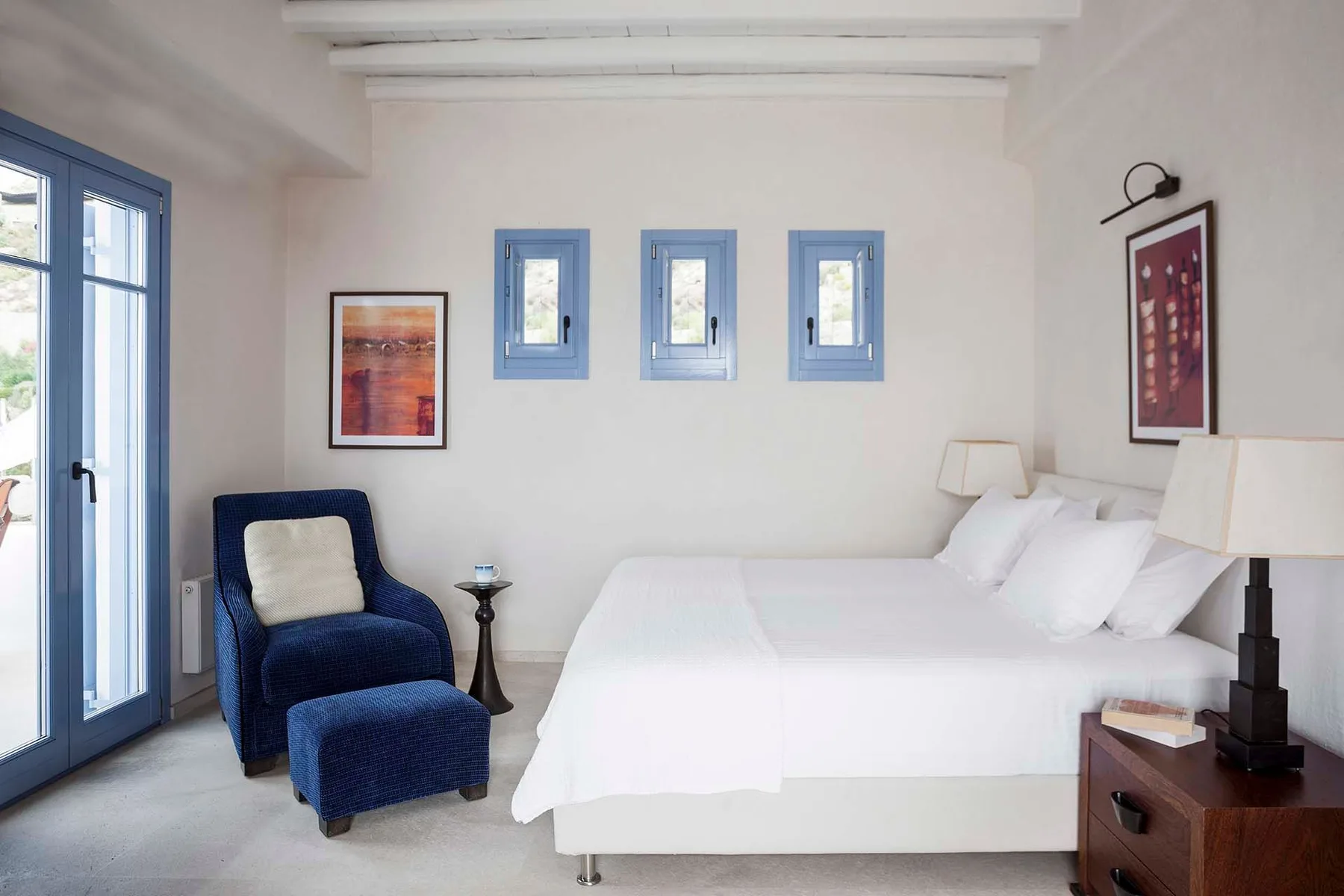 A bright bedroom with a white bed and blue seating beside glass doors.