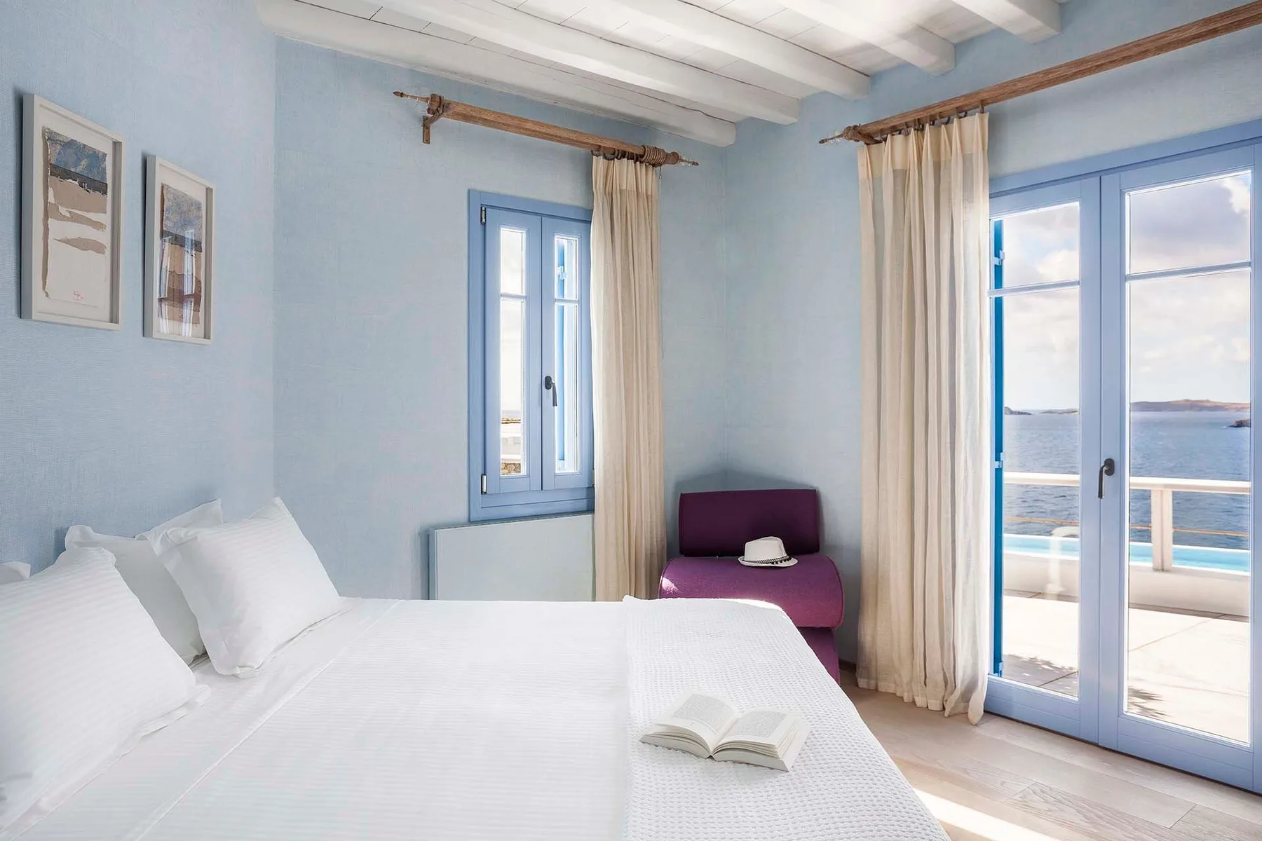 A bedroom with white bedding, blue-framed windows, and a view of the sea.