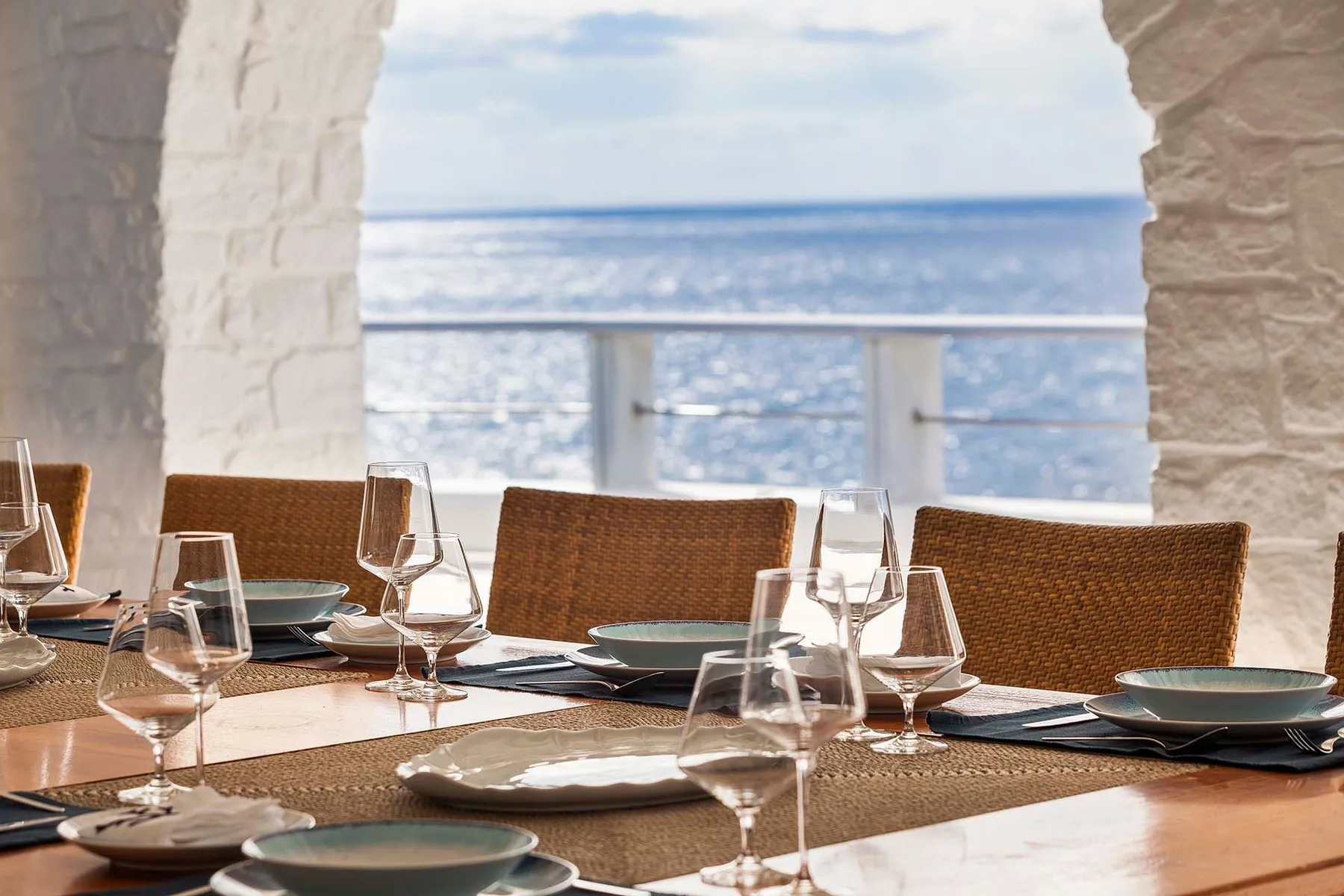 A seaside dining table is set with plates and glasses beside an arched opening.