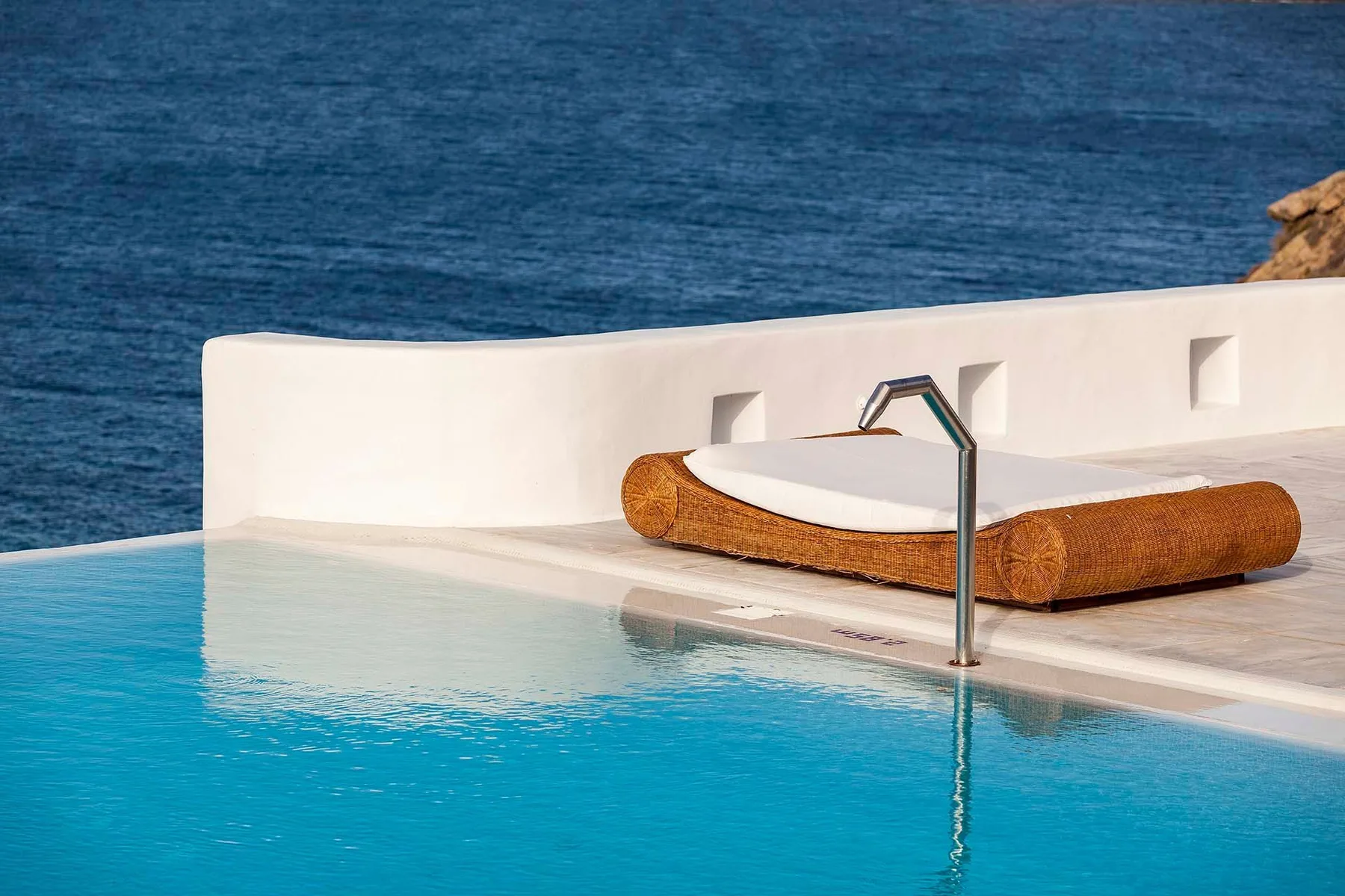 A swimming pool beside a sun deck with a low cushioned lounger overlooking the sea.