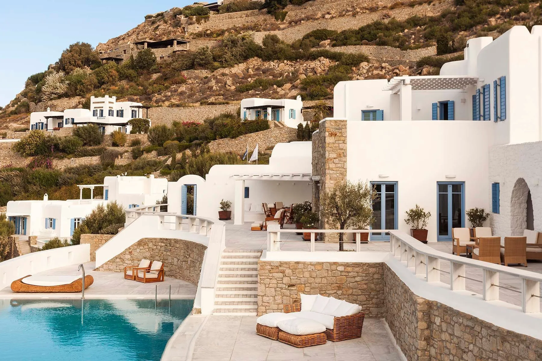 White hillside resort buildings overlook a turquoise pool and outdoor seating.