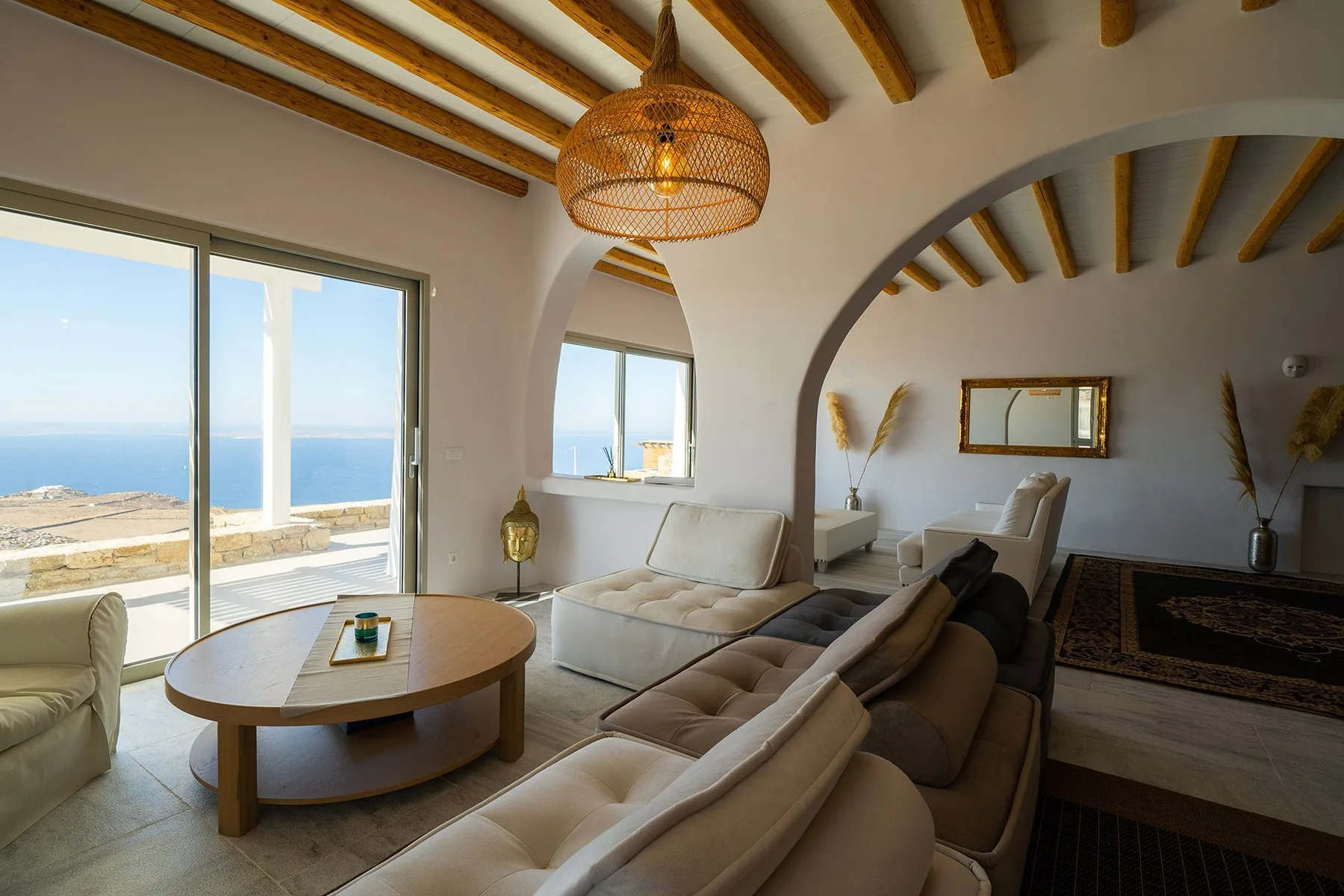 A bright living room with seating, a coffee table, and large windows overlooking the sea.