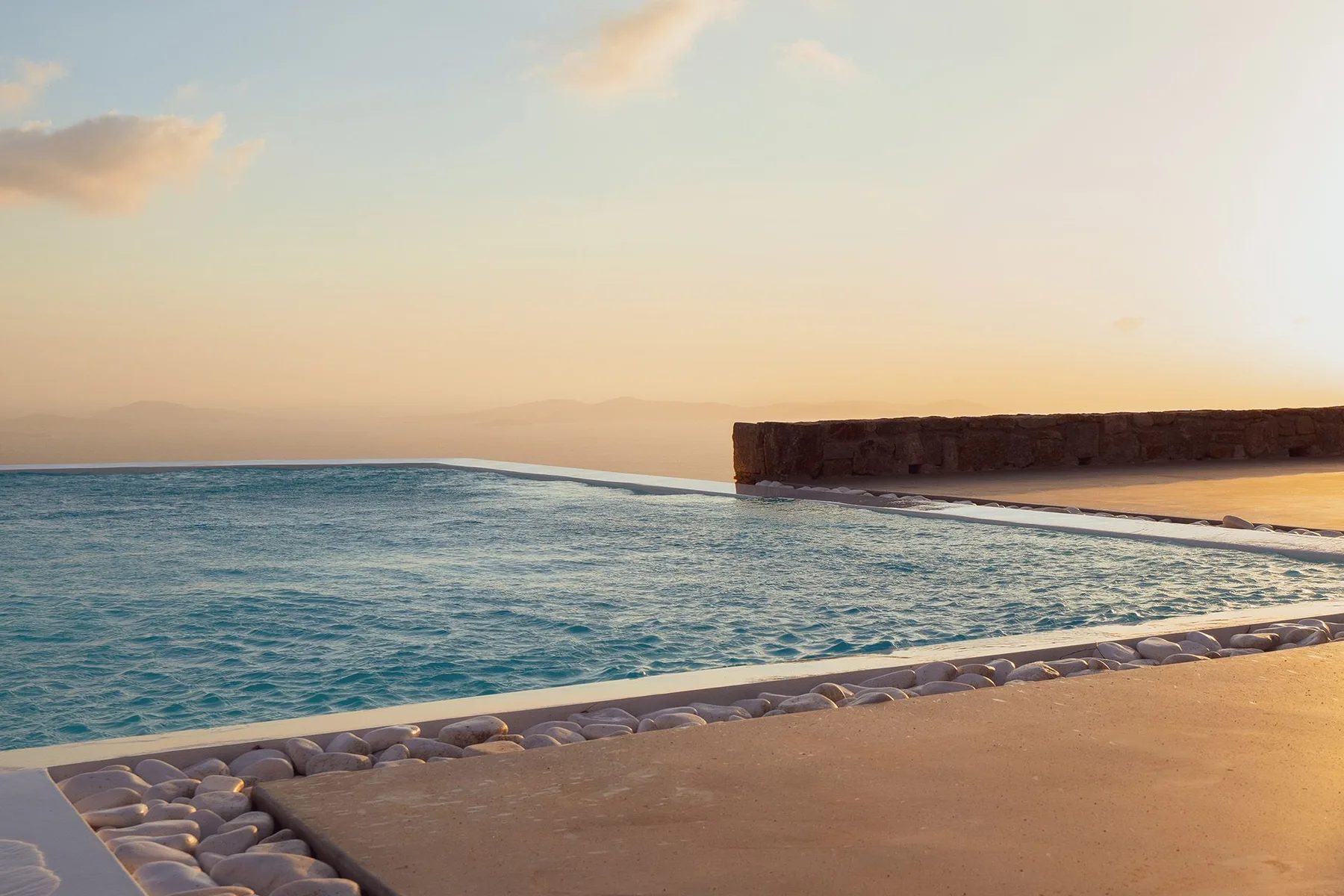 An outdoor swimming pool overlooks a low wall and distant hills at sunset.