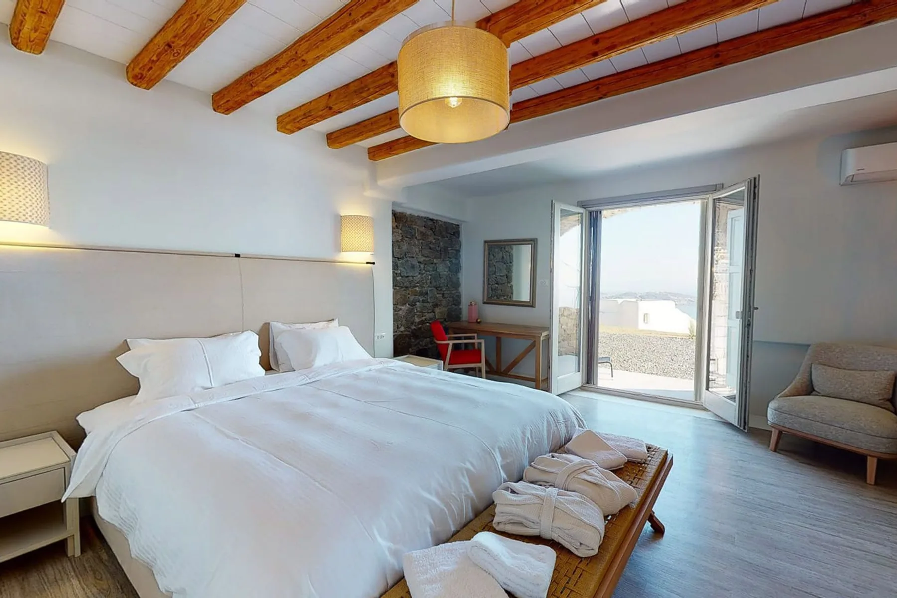 A bedroom with a large bed, exposed wooden ceiling beams, and open doors to a terrace.