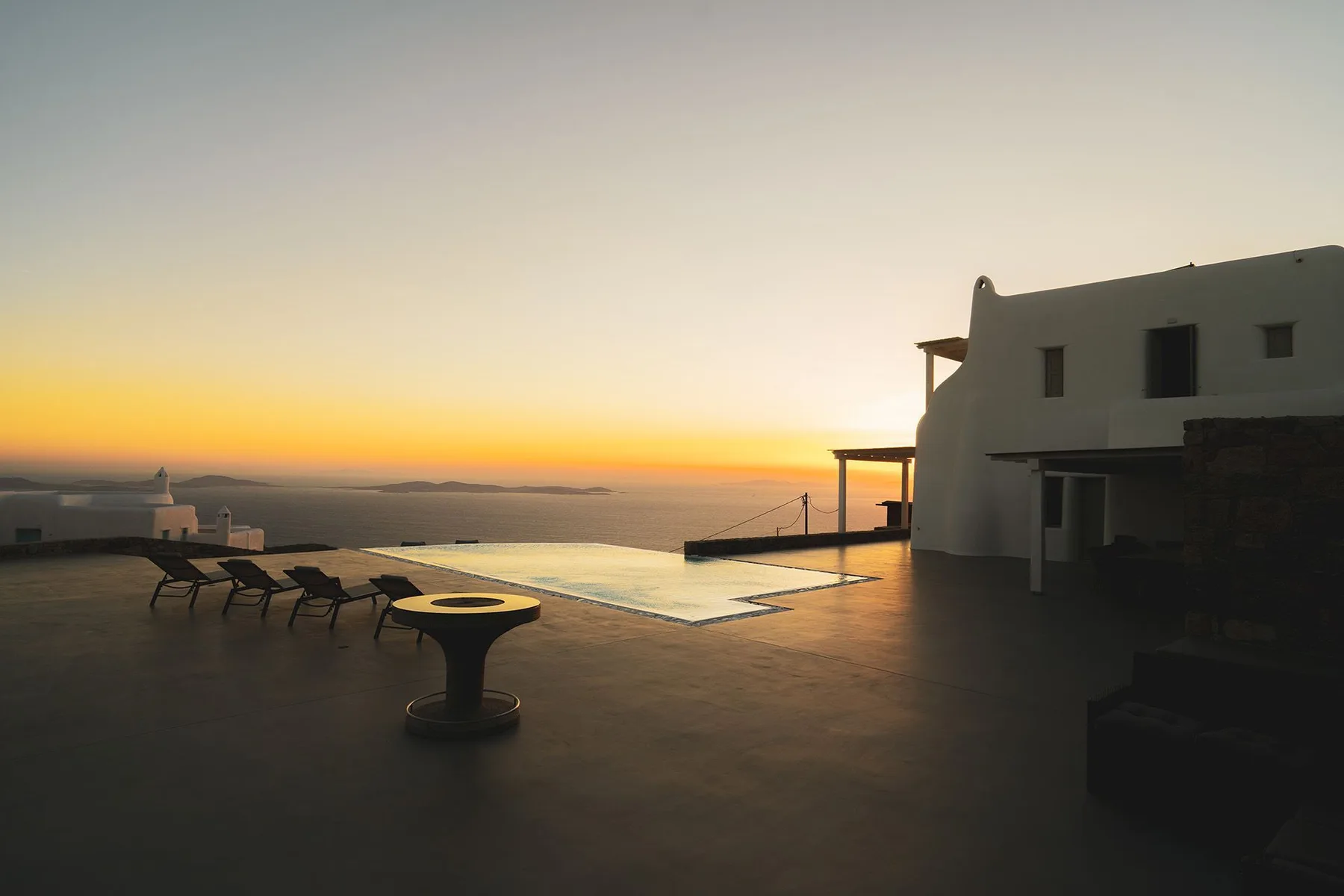 A rooftop terrace overlooks the sea at sunset, with lounge seating beside a pool.