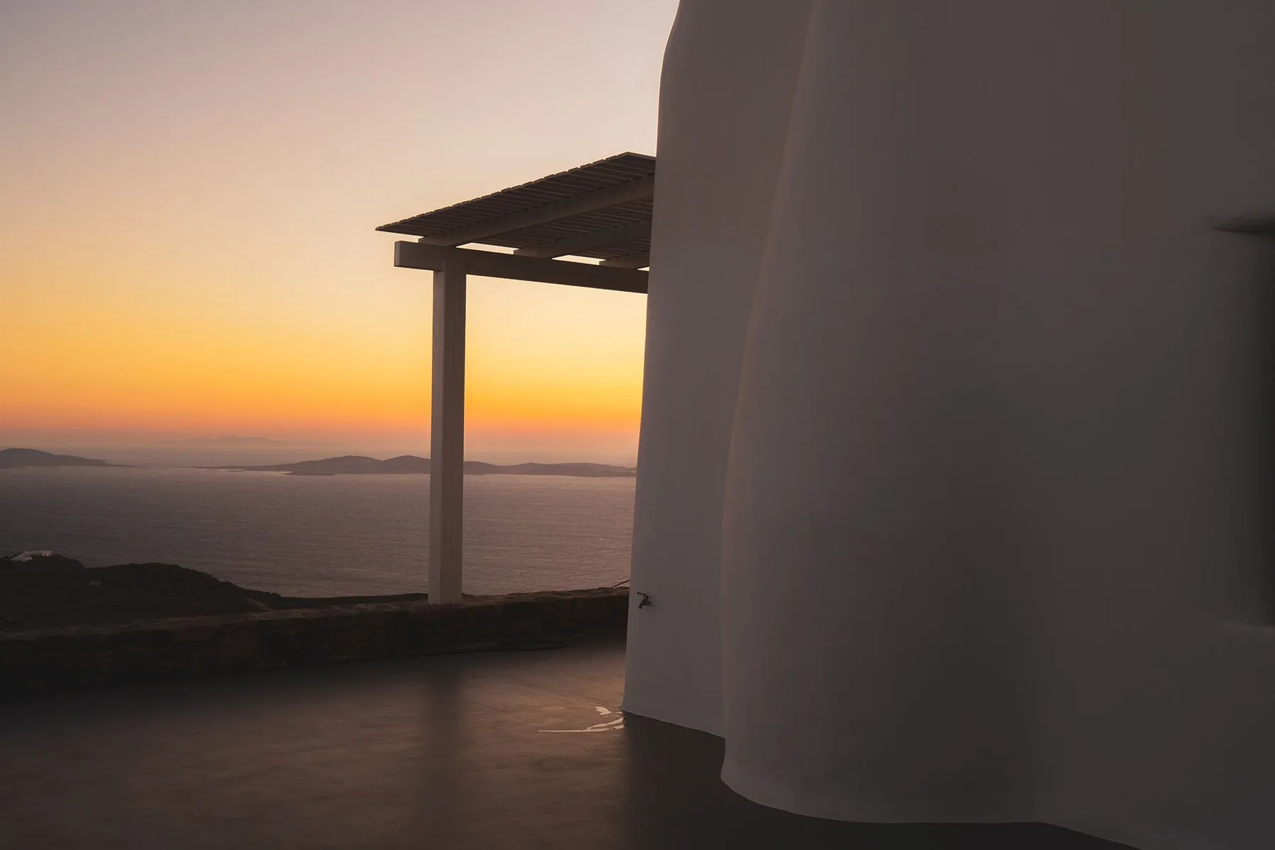 A terrace overlooks the sea at sunset beside a white curved wall.