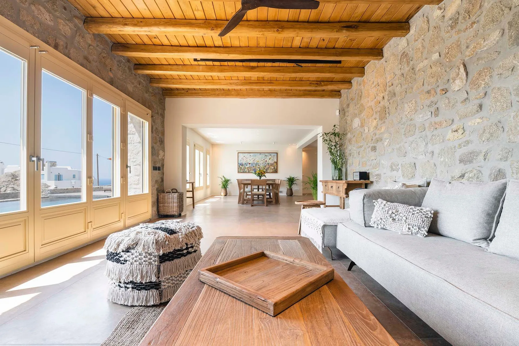 A bright living room with a seating area, wooden ceiling beams, and large windows.