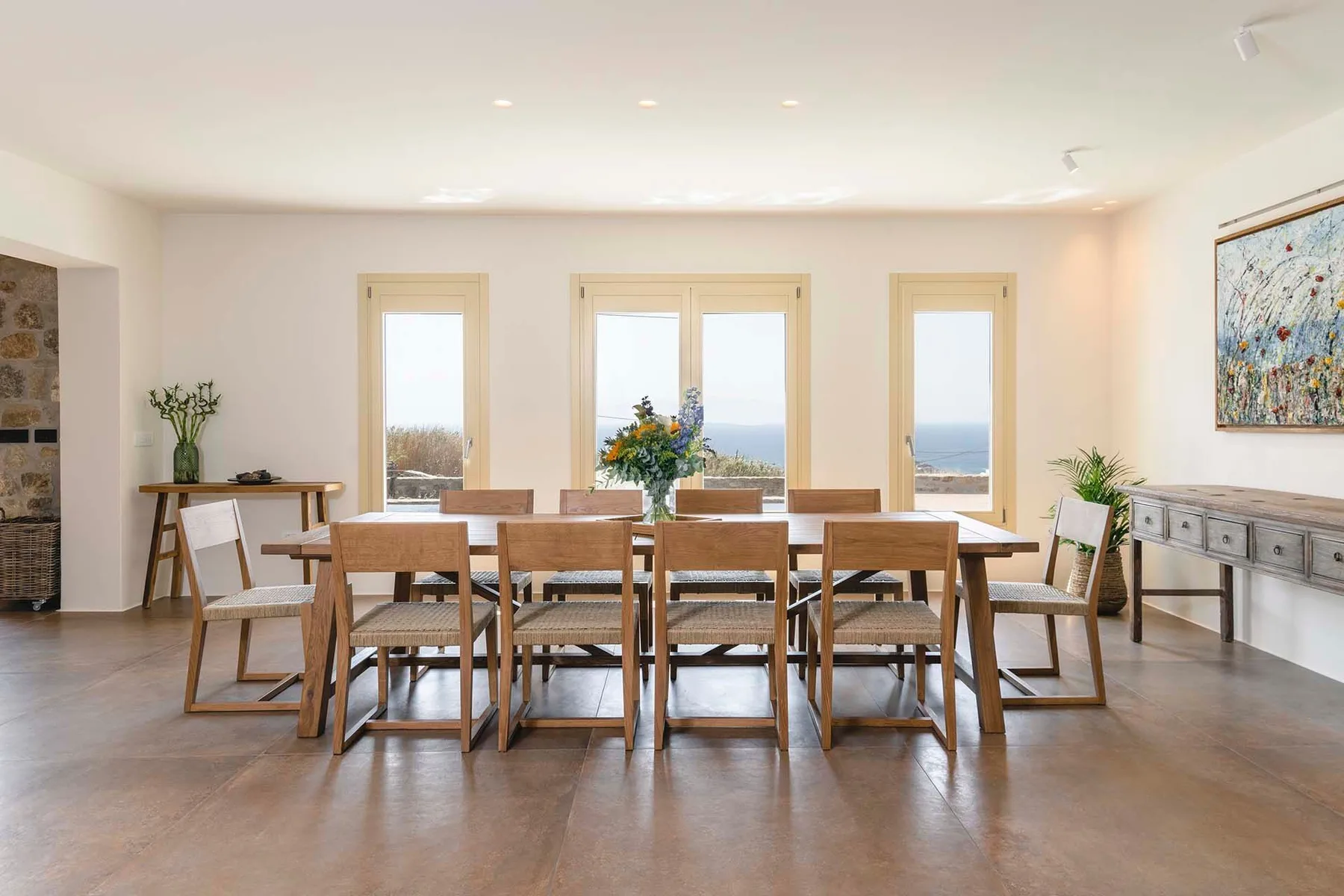 A bright dining room with a long wooden table and windows overlooking the water.