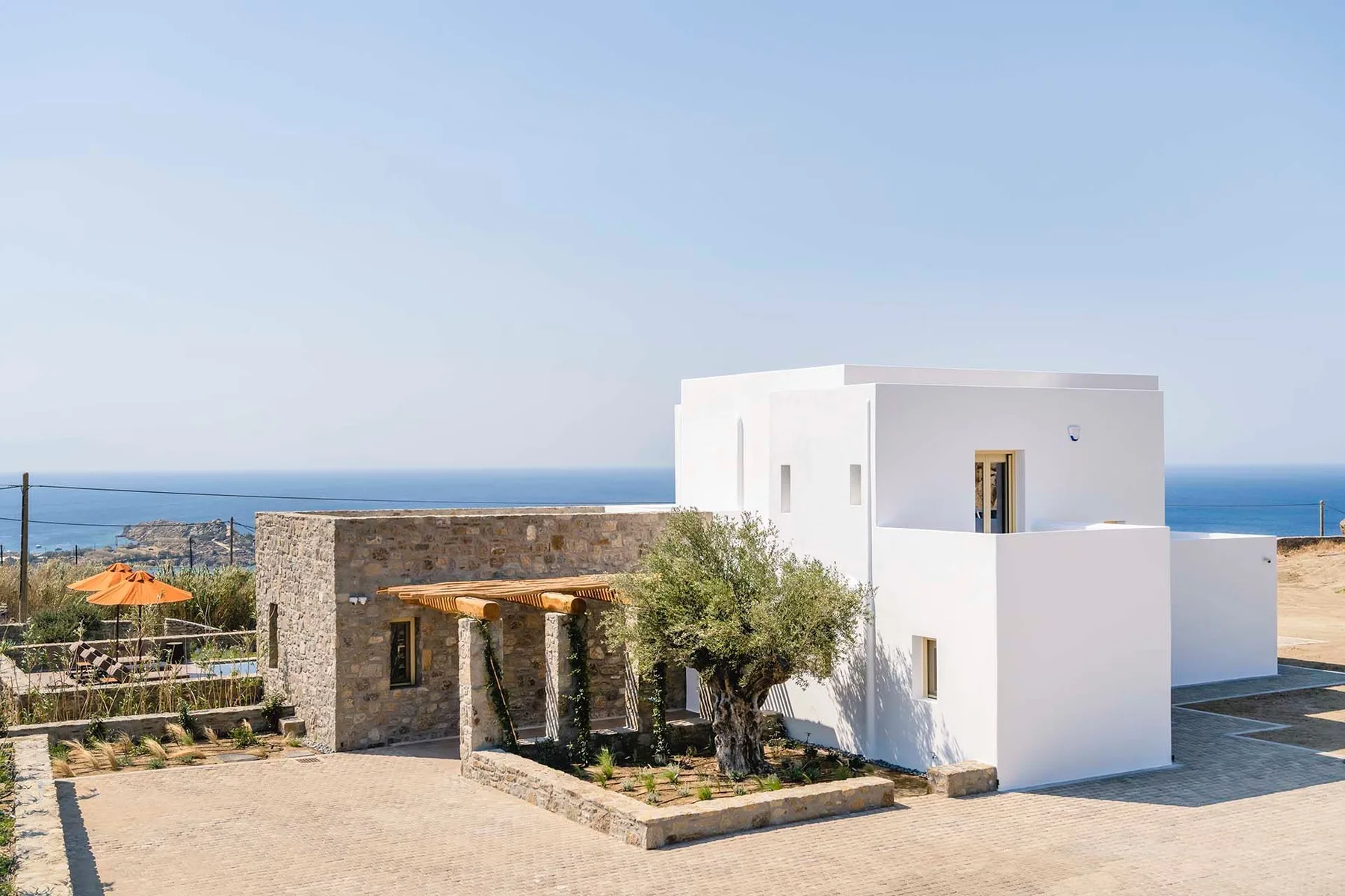 A modern white building and stone structure stand beside a paved courtyard overlooking the sea.
