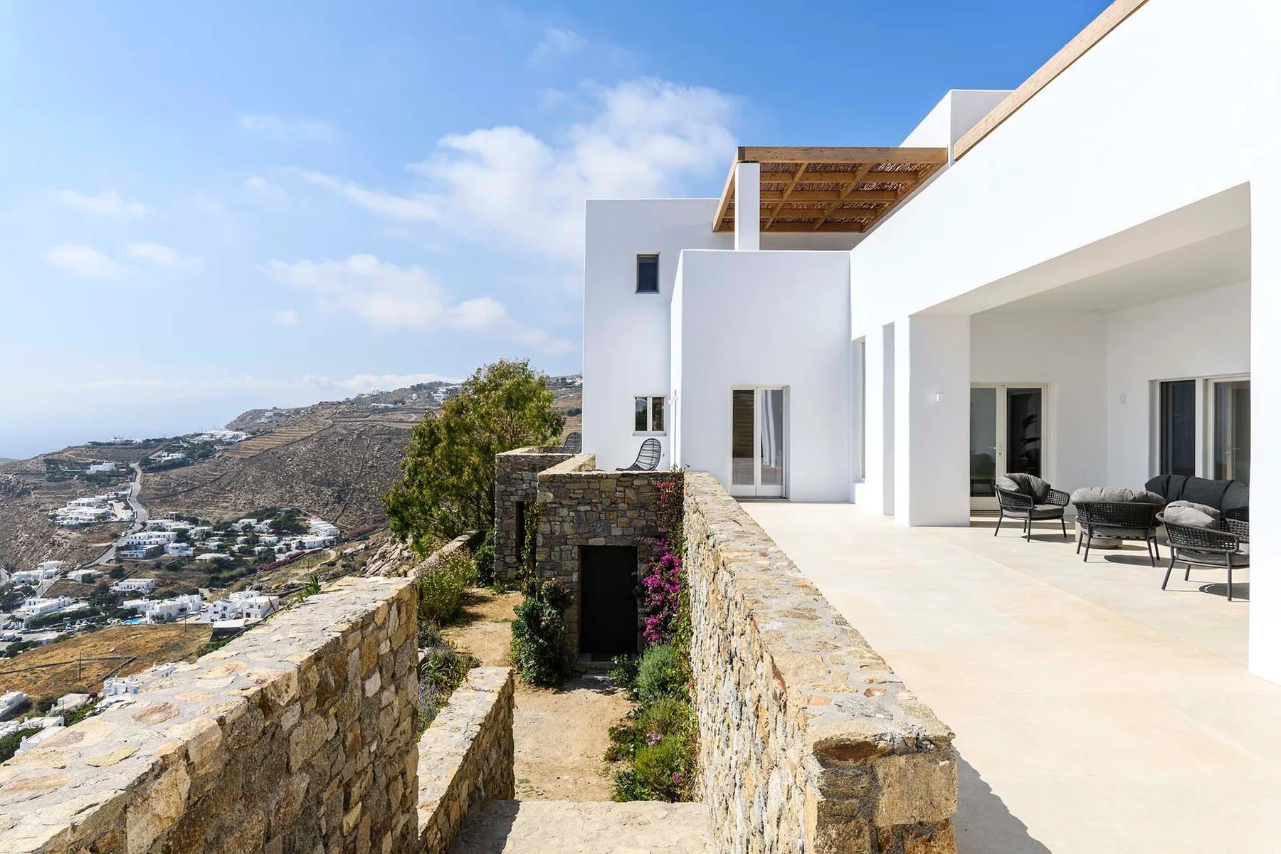 A modern white house opens onto a terrace overlooking a hilly landscape.