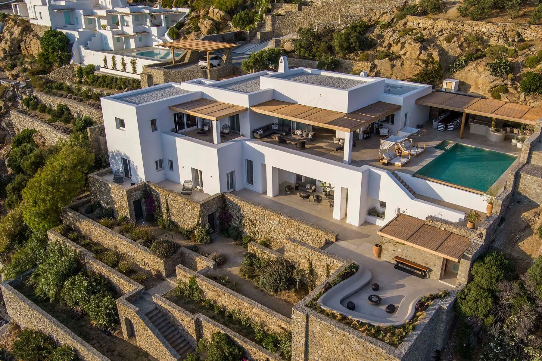 A modern white villa sits on a hillside beside a swimming pool and landscaped terraces.