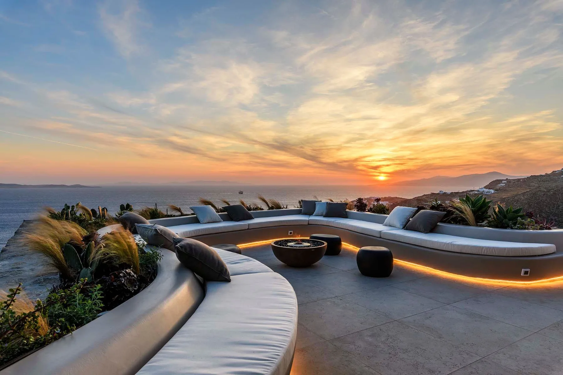 A curved outdoor lounge with cushioned seating overlooks the ocean at sunset.
