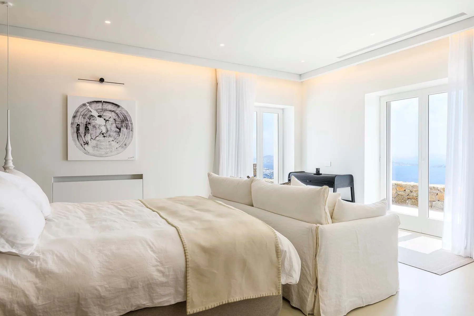 A bright bedroom with a large bed, seating, and tall windows overlooking the water.
