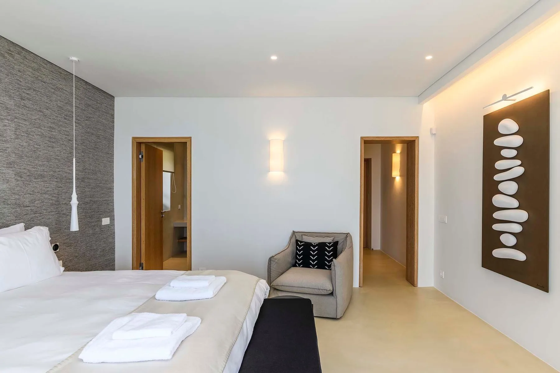 A bright bedroom with a bed, a cushioned chair, and framed wall art.
