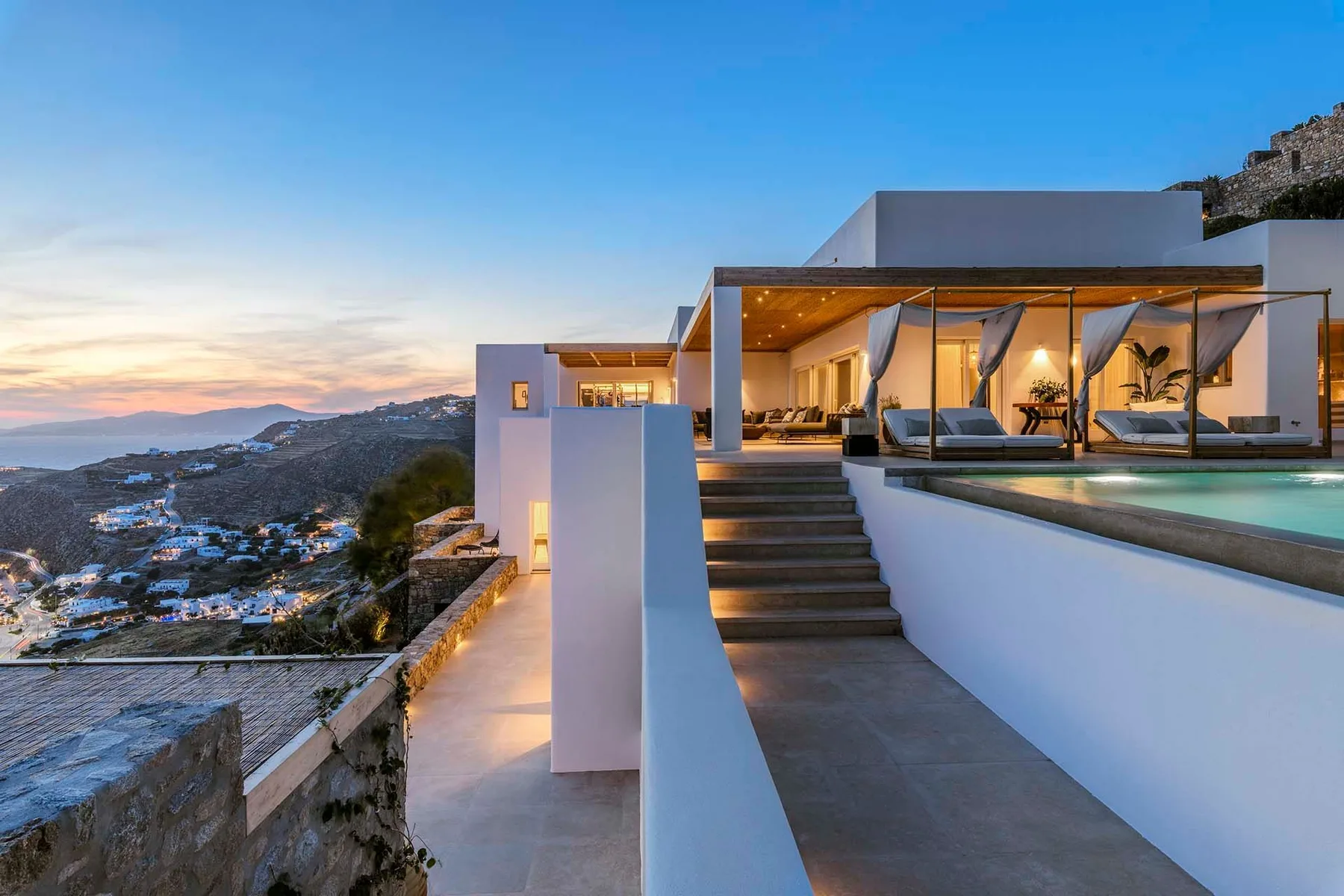 A modern hillside house opens onto a terrace with seating overlooking the coast at sunset.