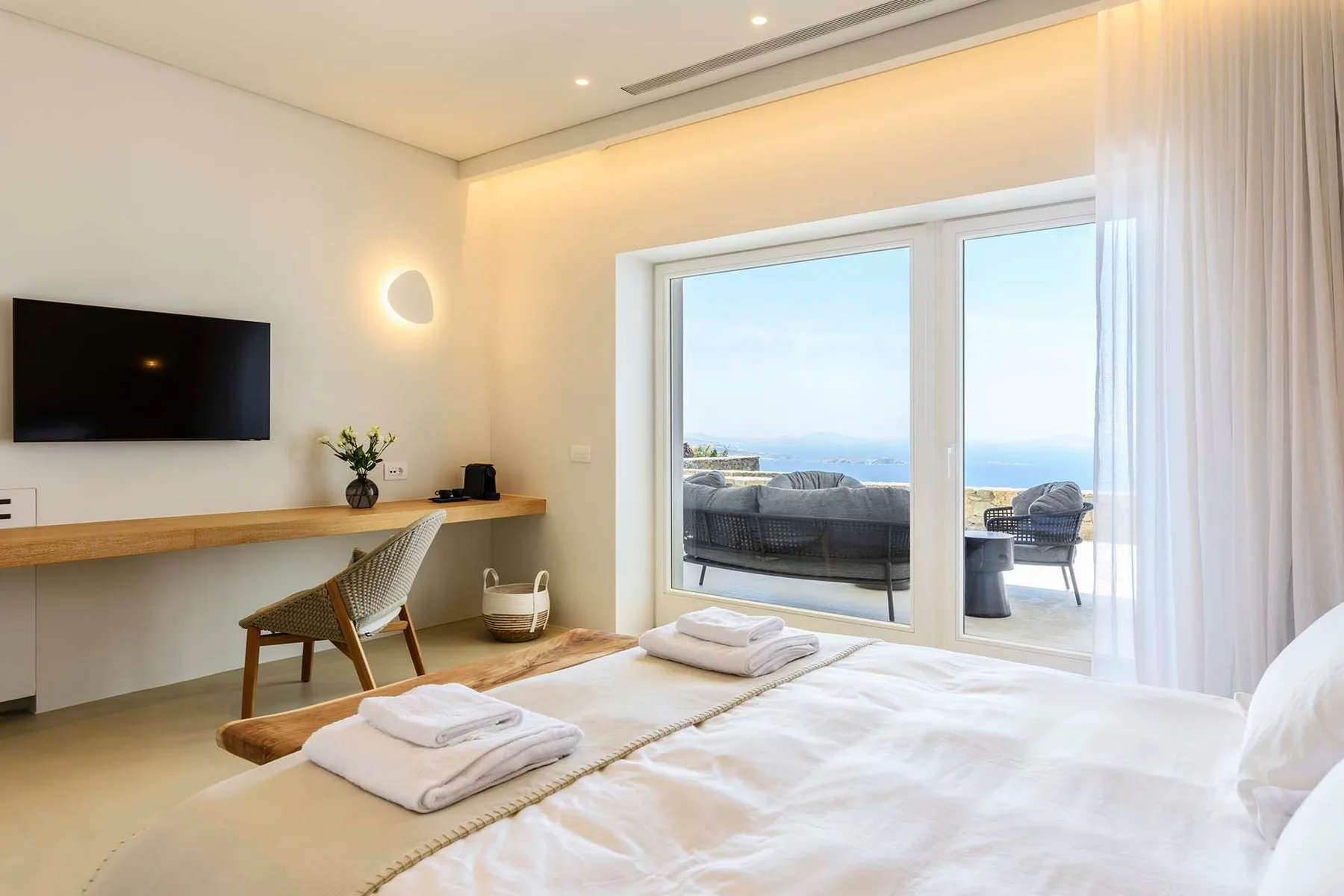 A bright bedroom with a desk, television, and glass doors opening onto a sea view.
