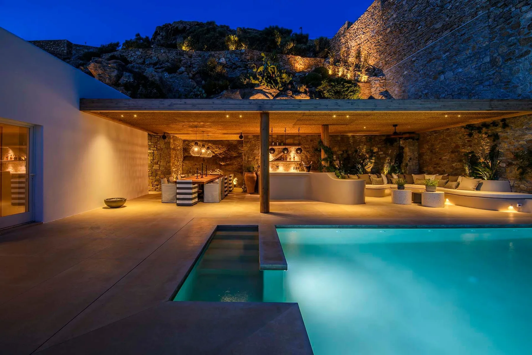 An illuminated outdoor pool beside a covered patio and stone walls at dusk.