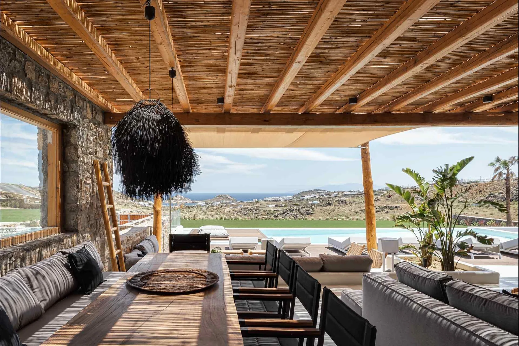Una sala exterior cubierta se abre a un paisaje luminoso, con asientos acolchados y una mesa larga.