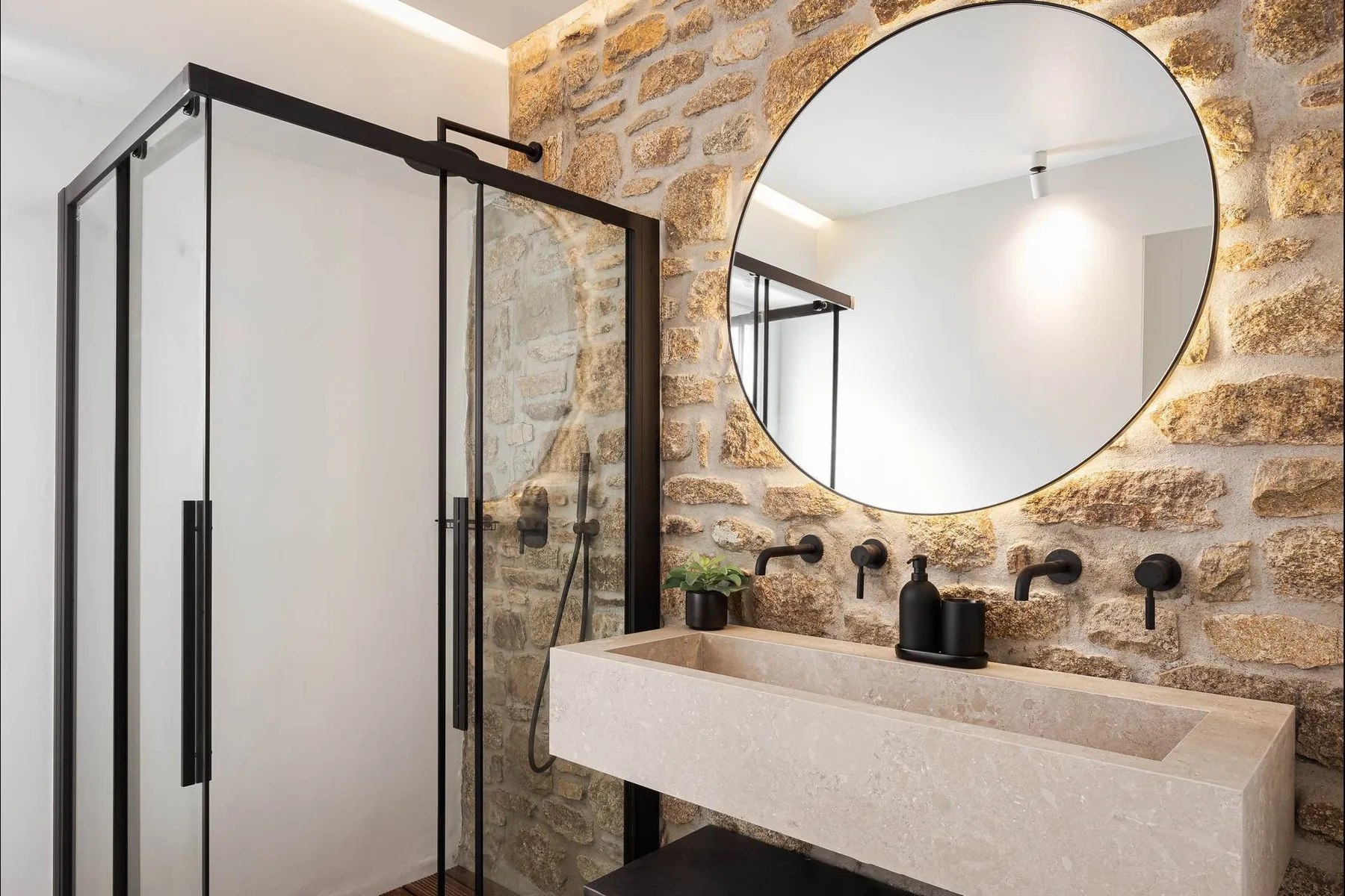 Un baño con ducha de vidrio, paredes de piedra y un tocador largo bajo un espejo redondo.