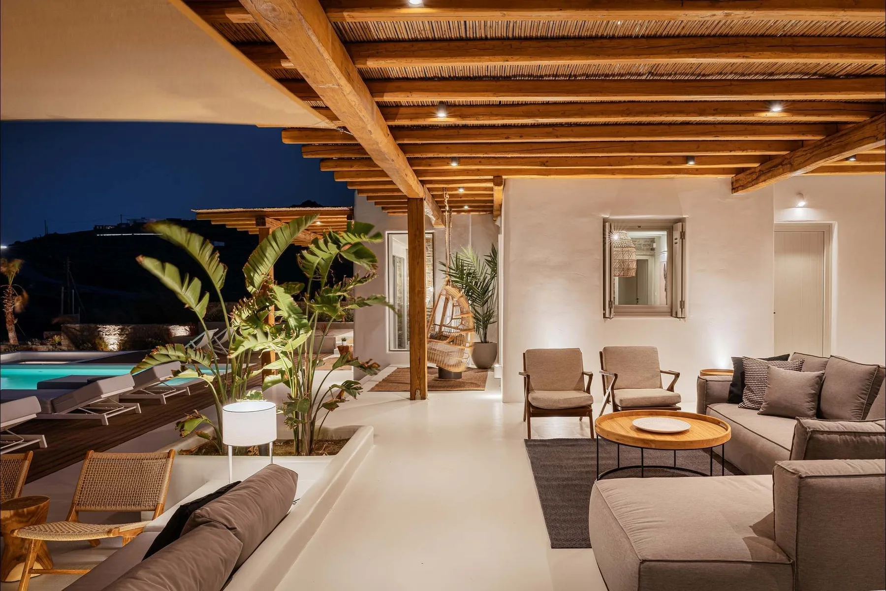 Una zona de estar exterior cubierta da a una piscina, con muebles de descanso y plantas.