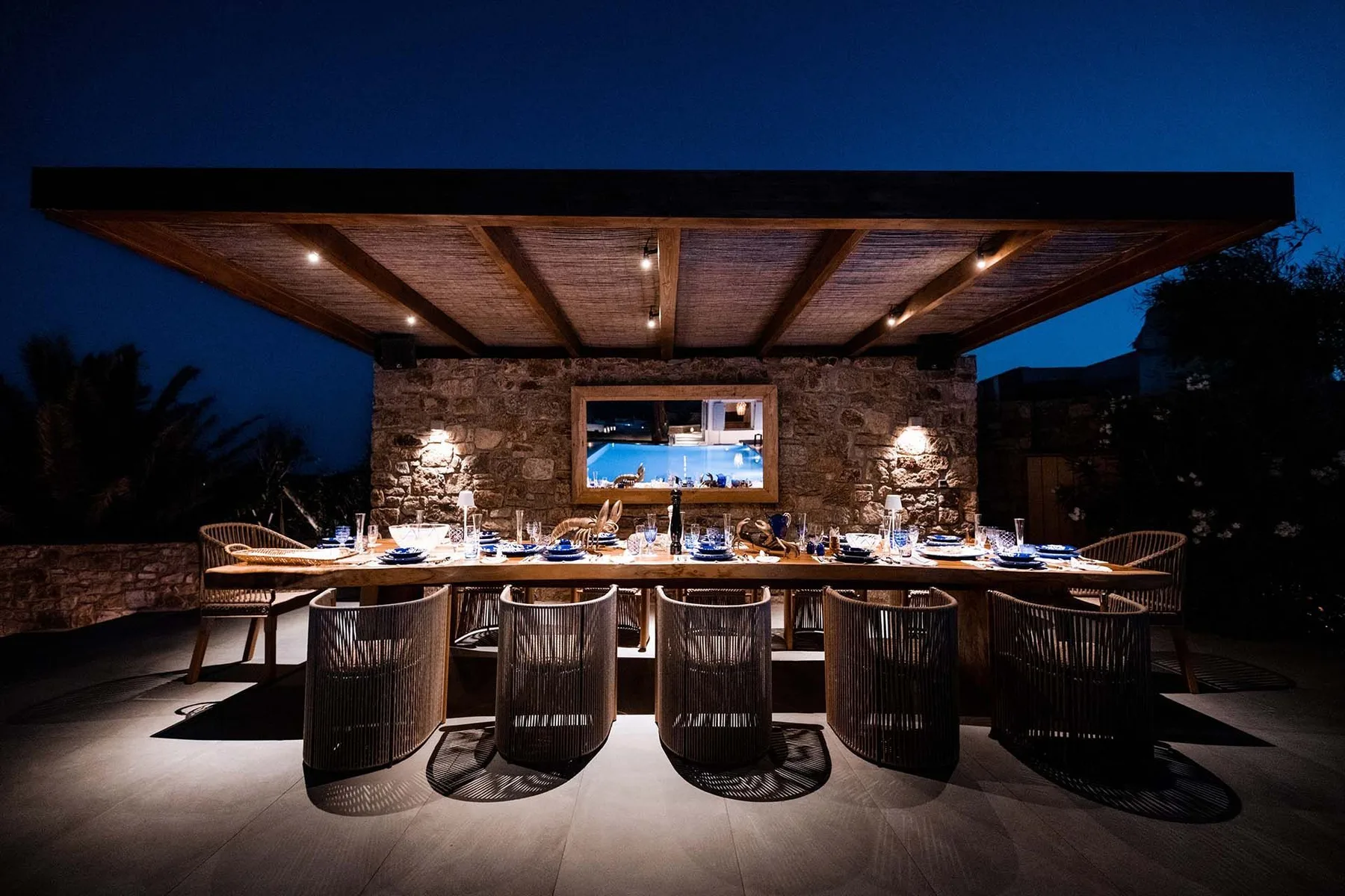 An outdoor dining area under a wooden canopy, with a long table set for a meal.