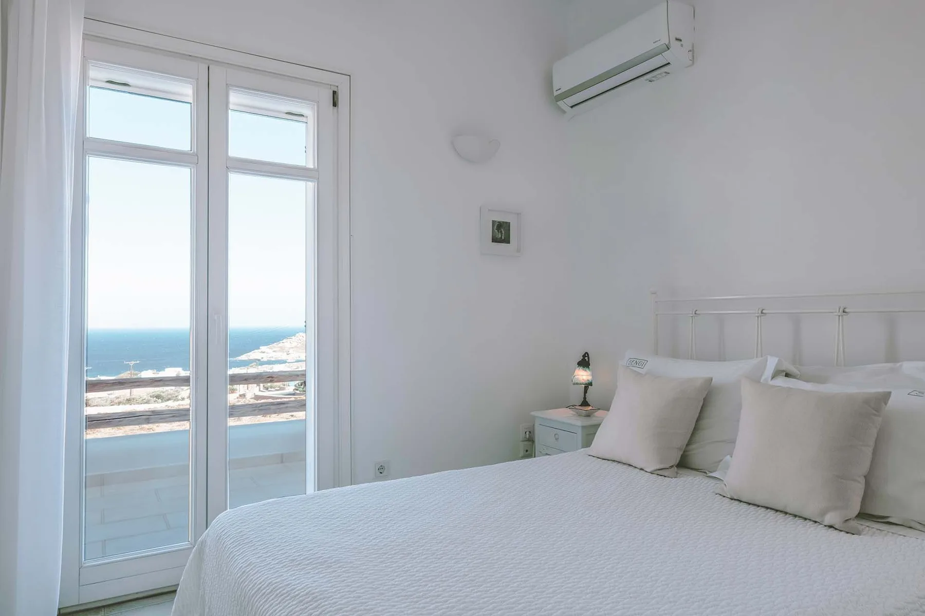 A bedroom with a bed beside tall windows overlooking the sea.