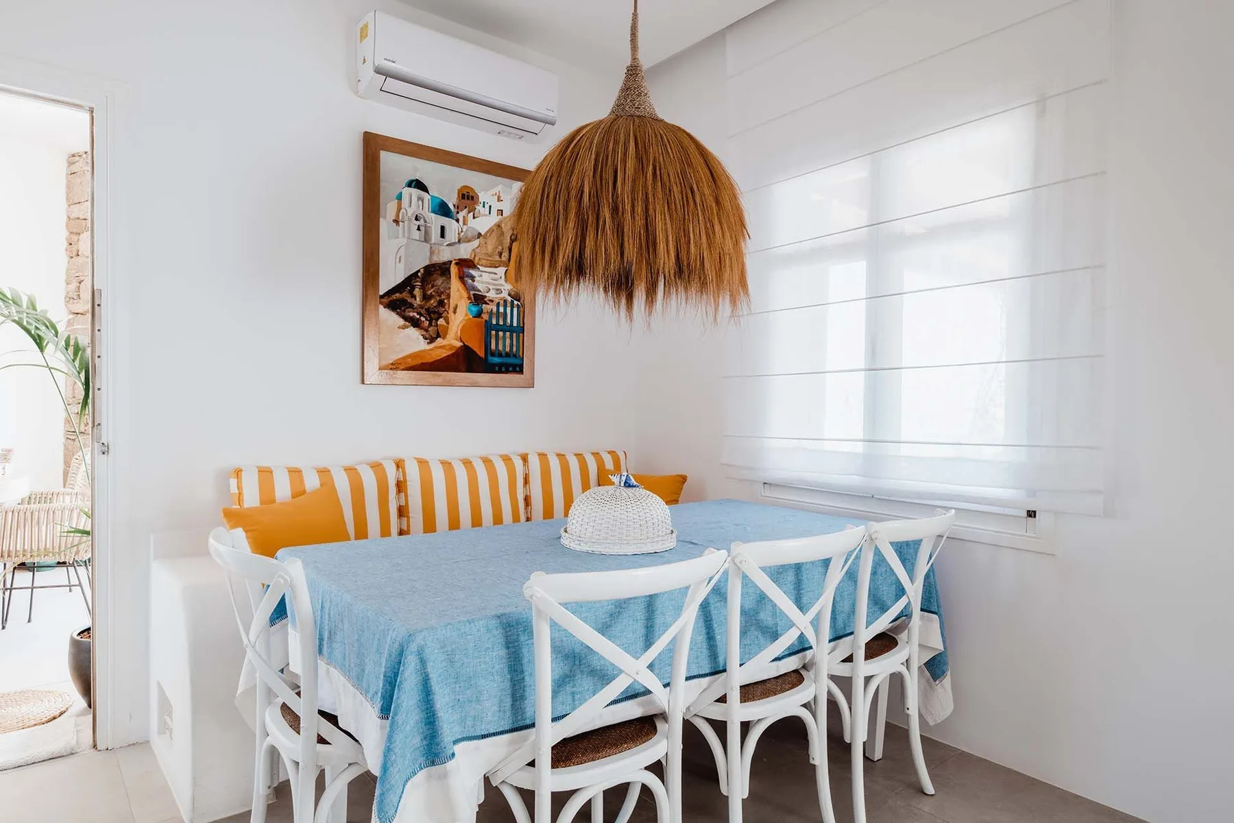 A bright dining area with a long table, white chairs, and a woven pendant light.