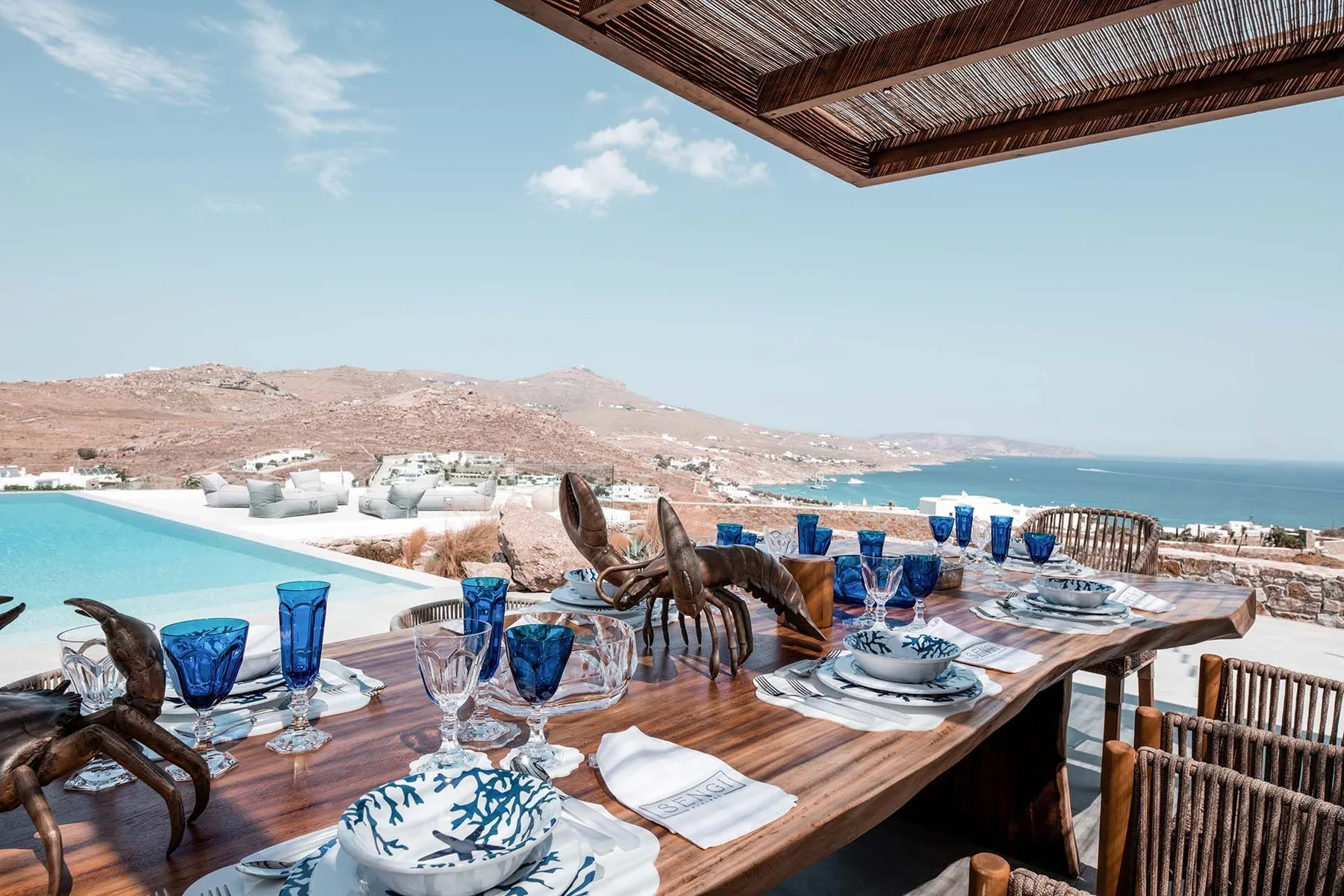 An outdoor dining terrace overlooks a pool and the sea, with a table set for a meal.