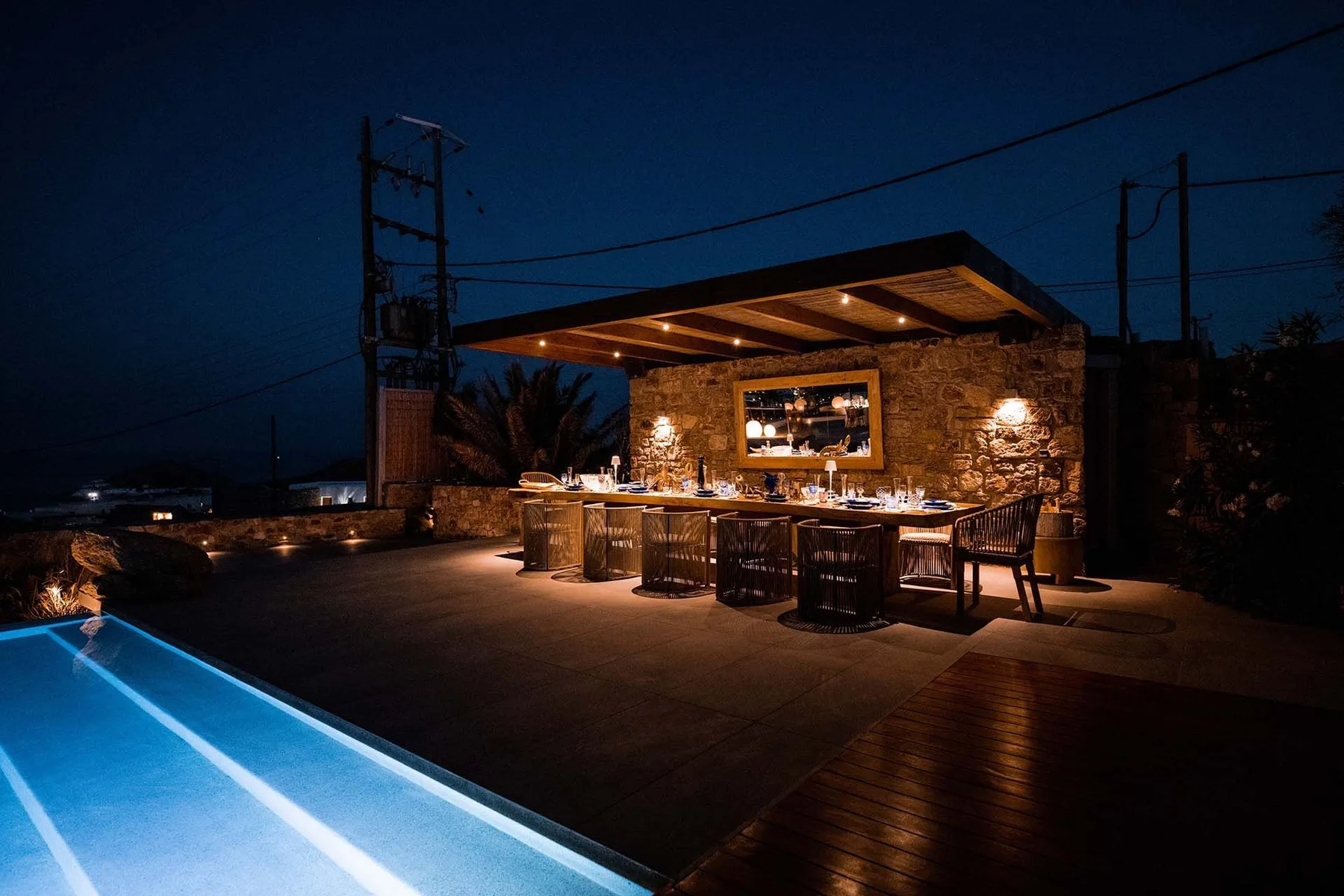 An outdoor dining area glows beneath a covered stone counter beside a swimming pool at night.