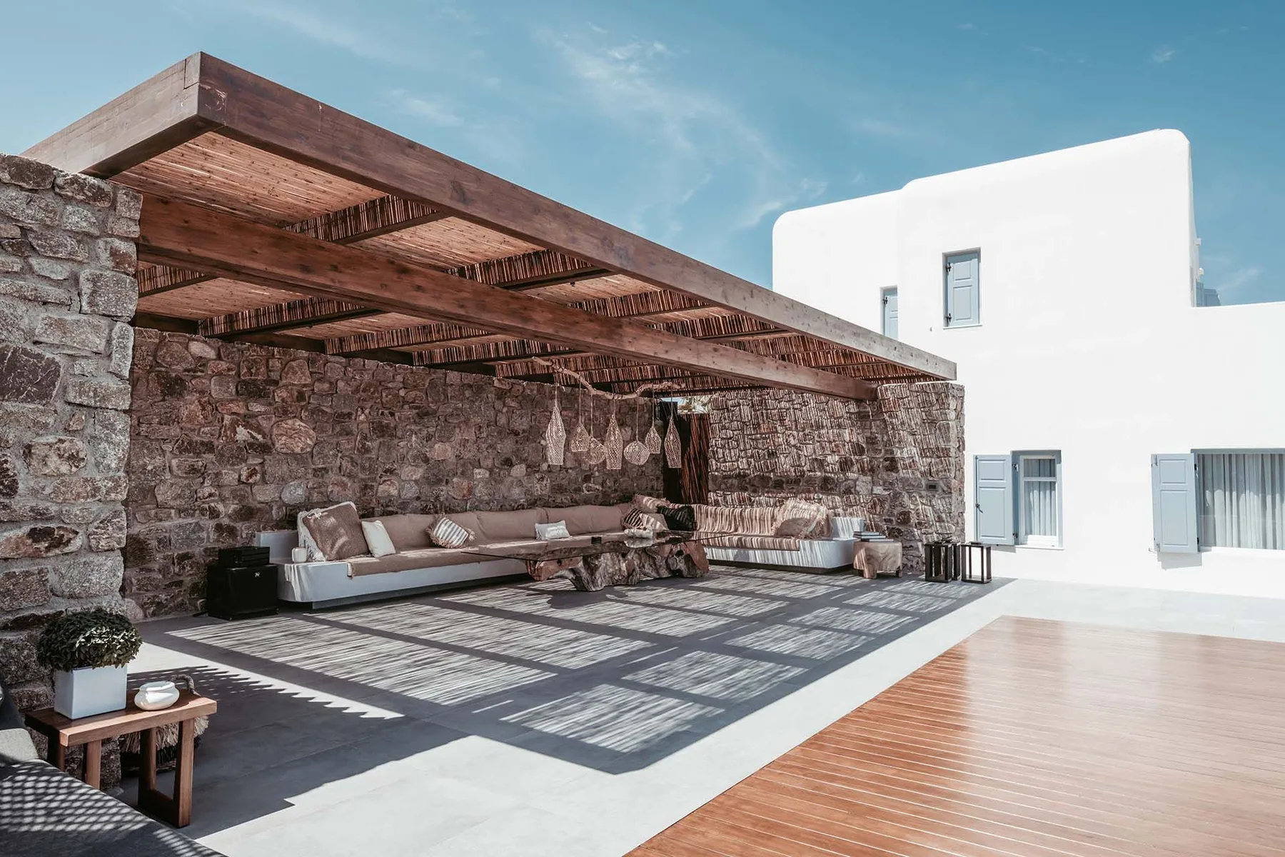 An outdoor lounge area with seating beneath a wooden pergola beside a white building.