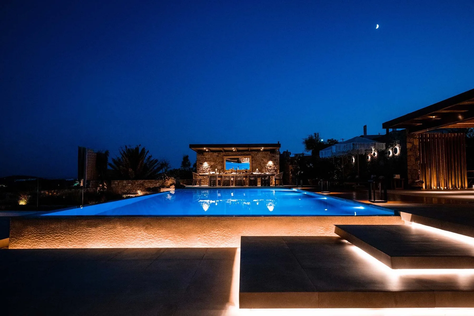 An illuminated outdoor swimming pool at night, with a lit building in the background.