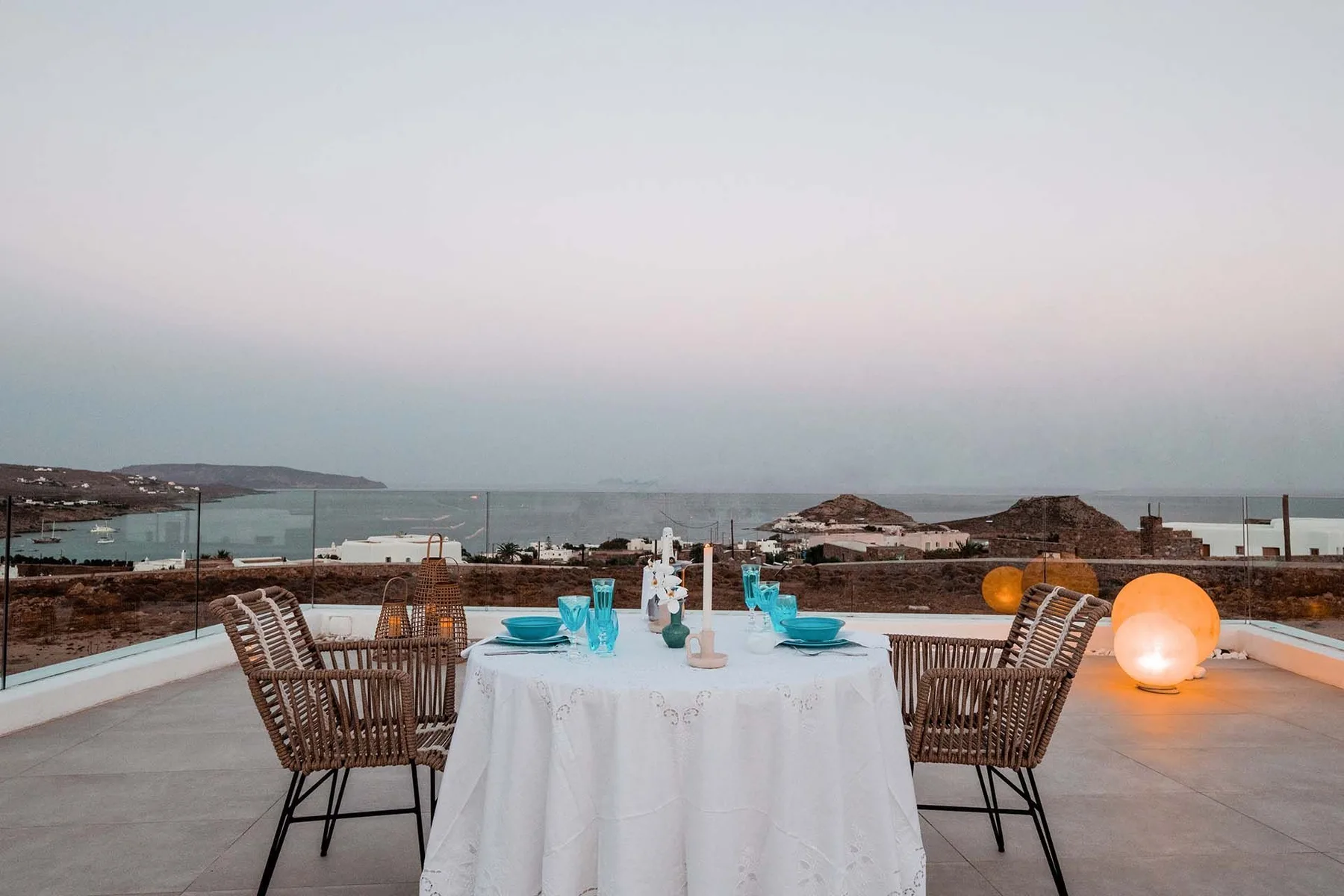 A rooftop dining area has a table set with dishes and chairs, overlooking the sea.