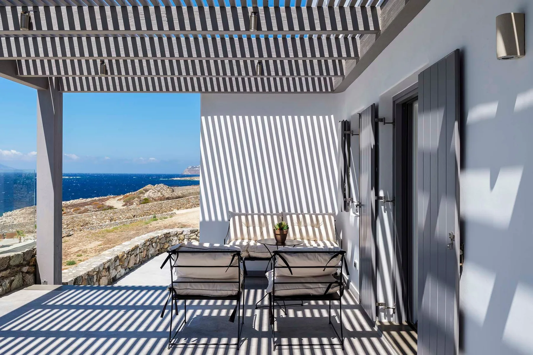 A shaded seaside terrace has a small table and seating overlooking the water.