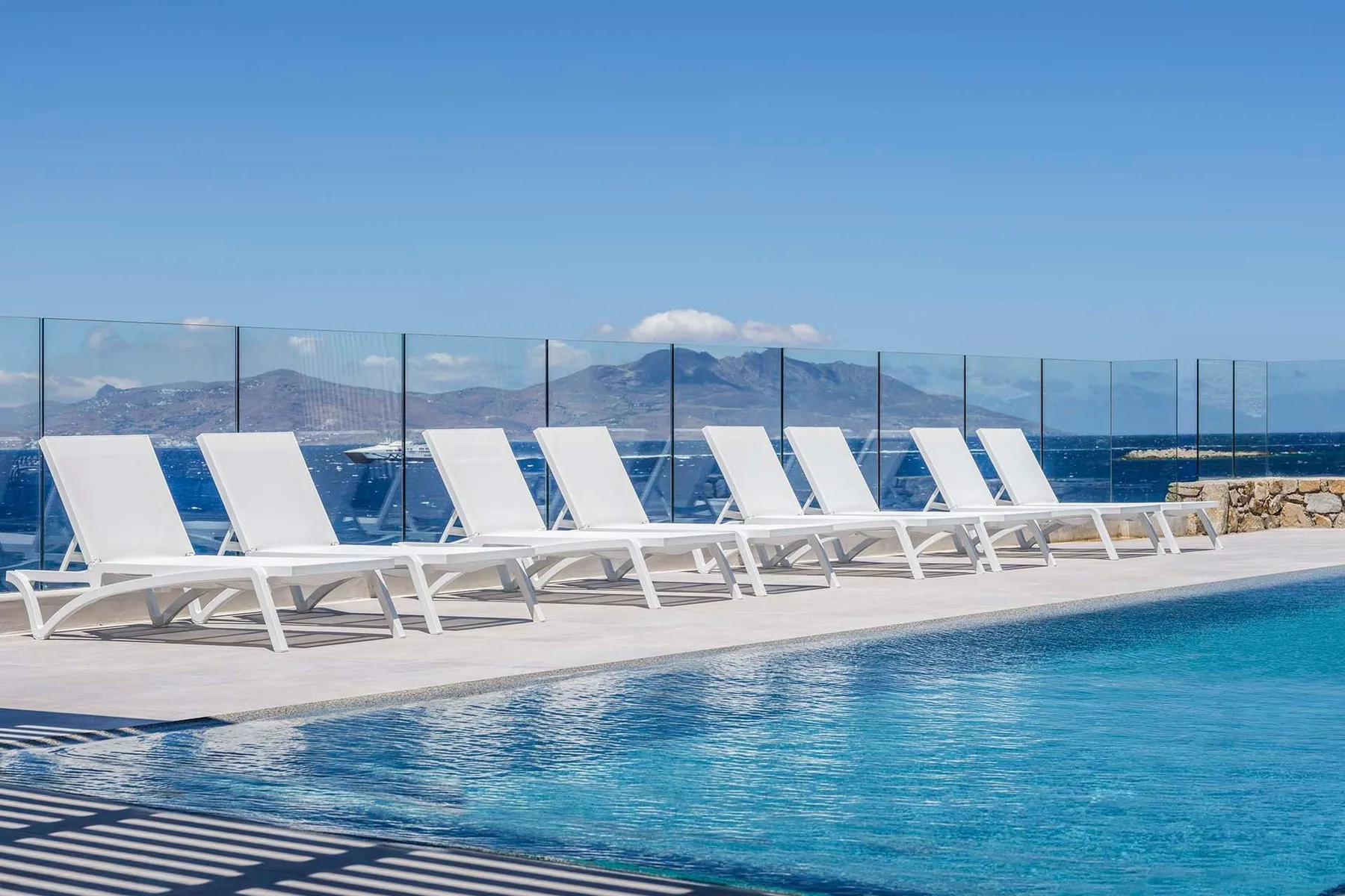 A swimming pool beside a row of loungers, with the sea and mountains beyond.