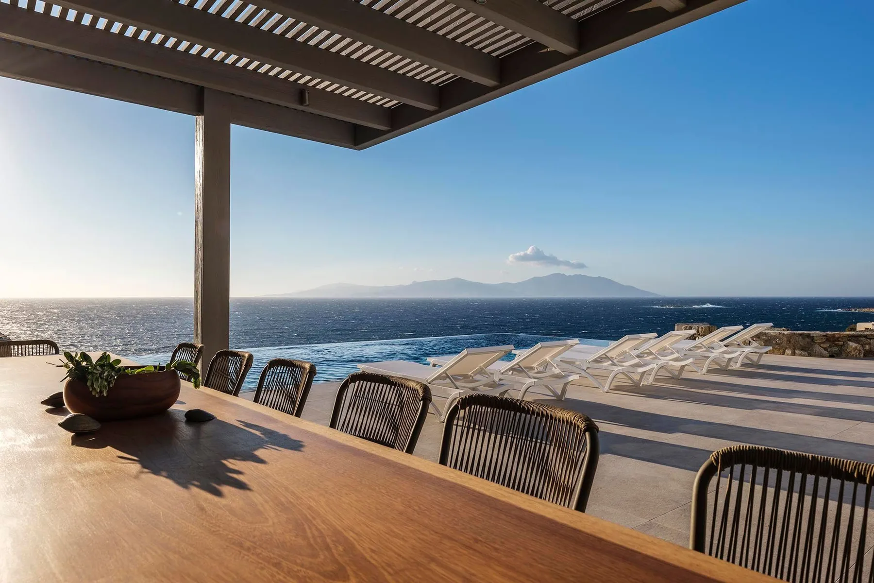A seaside terrace with a wooden dining table, lounge seating, and views across the water.