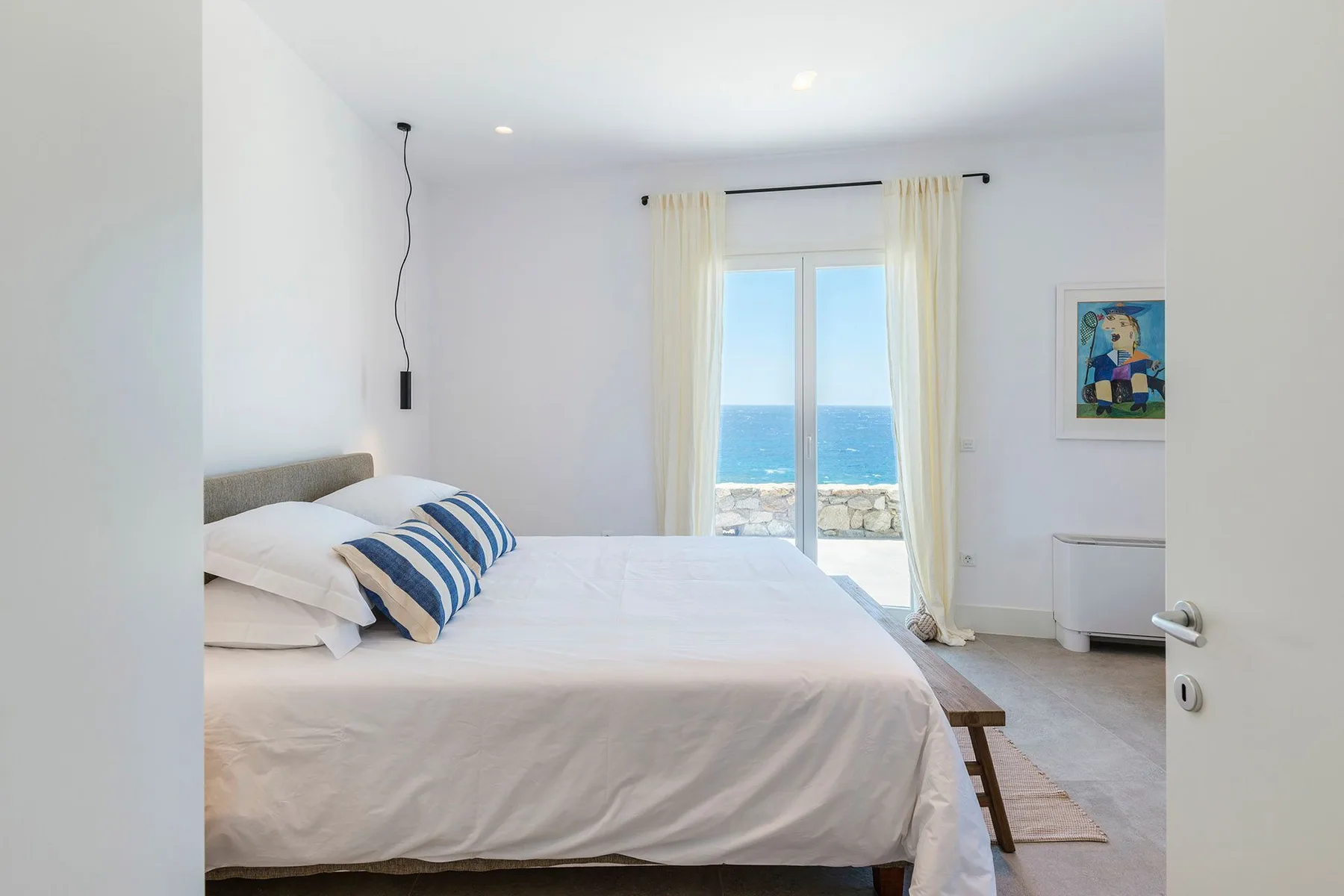 A bright bedroom with a bed facing a window overlooking the sea.
