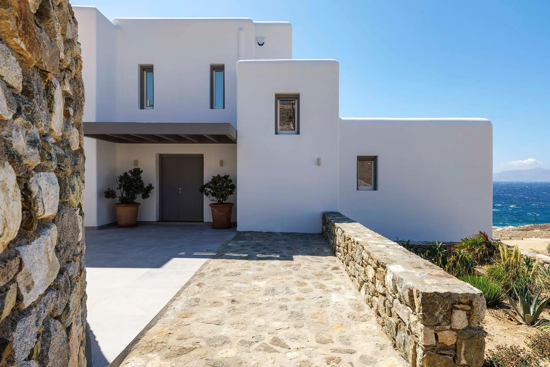 A white modern building with a stone-paved entrance and the sea visible nearby.