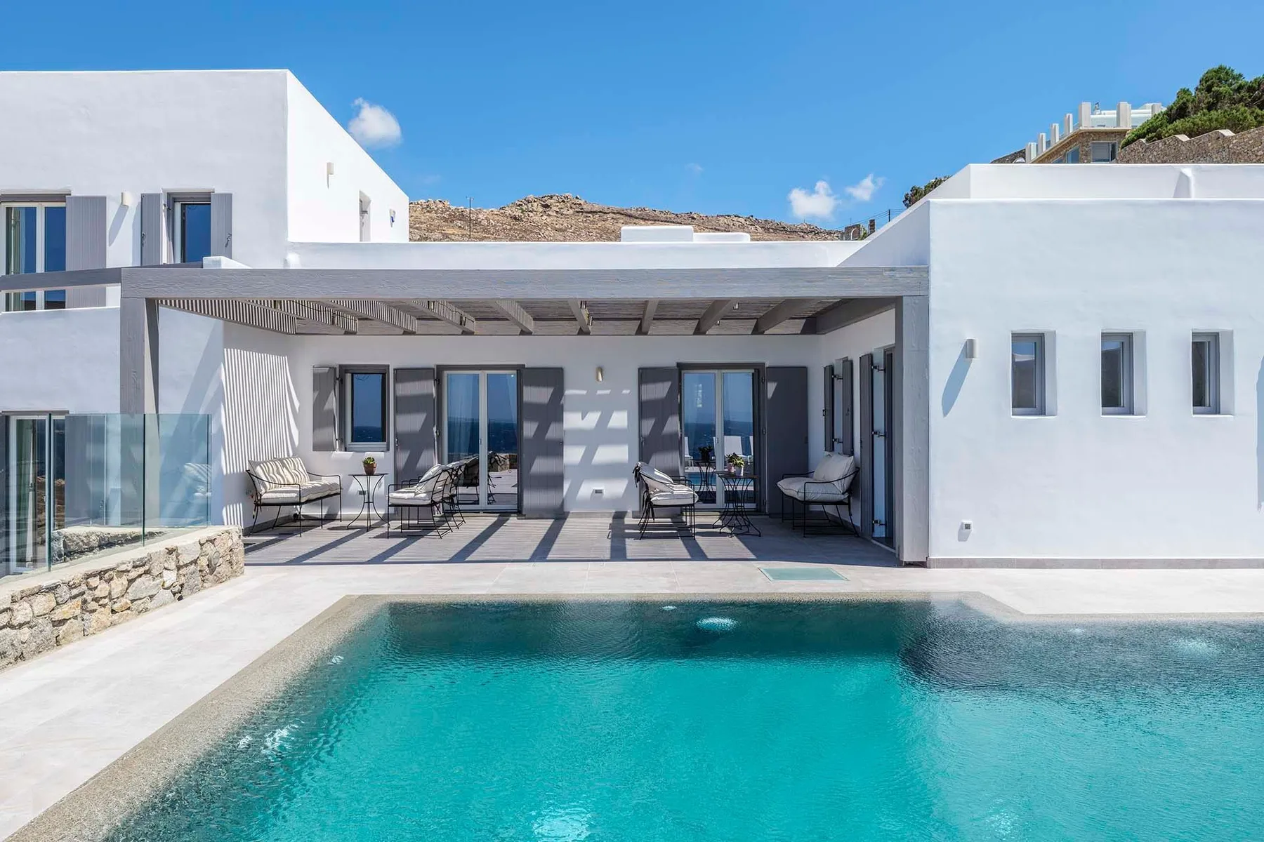 A modern white house overlooks a swimming pool, with shaded seating beneath a covered patio.