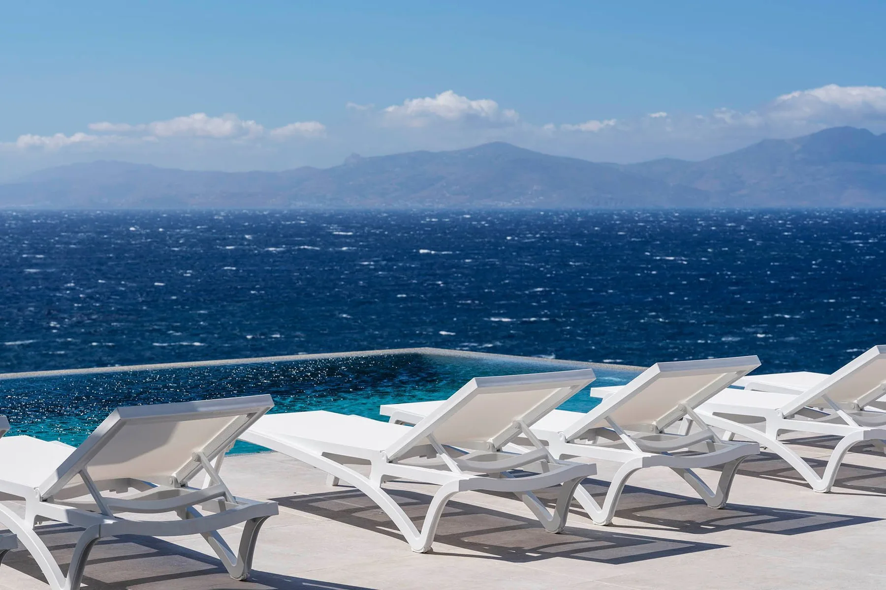 White lounge chairs line a poolside terrace overlooking the sea and distant mountains.