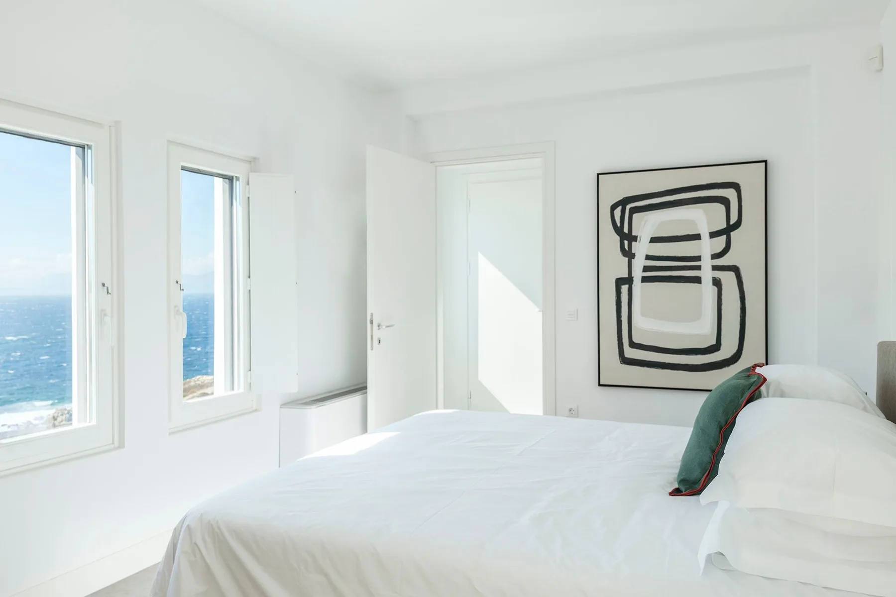 A bright white bedroom with a bed, abstract wall art, and windows overlooking the sea.