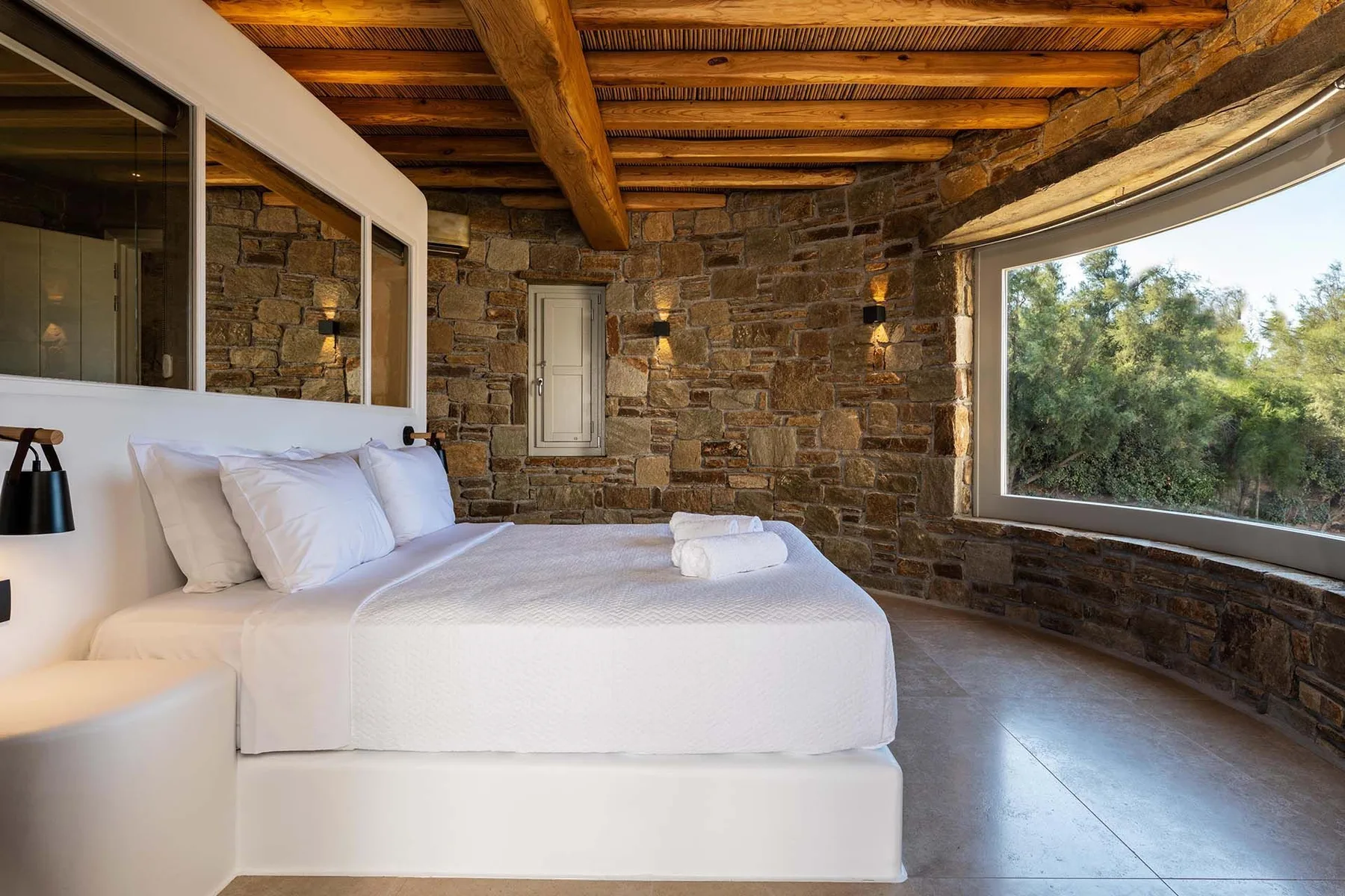 A bedroom with a white bed beside a large window overlooking greenery.