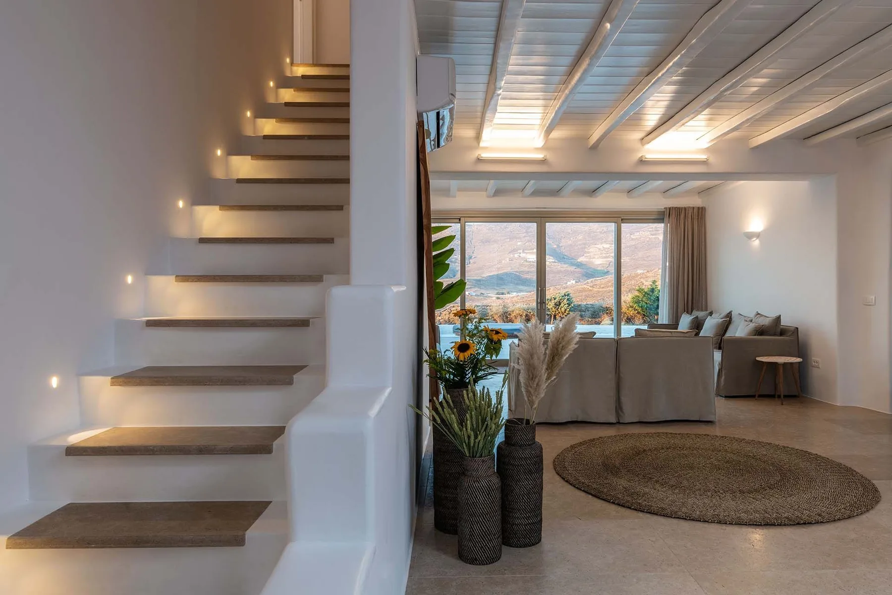 A bright living room with a staircase, seating, and large windows overlooking the hills.