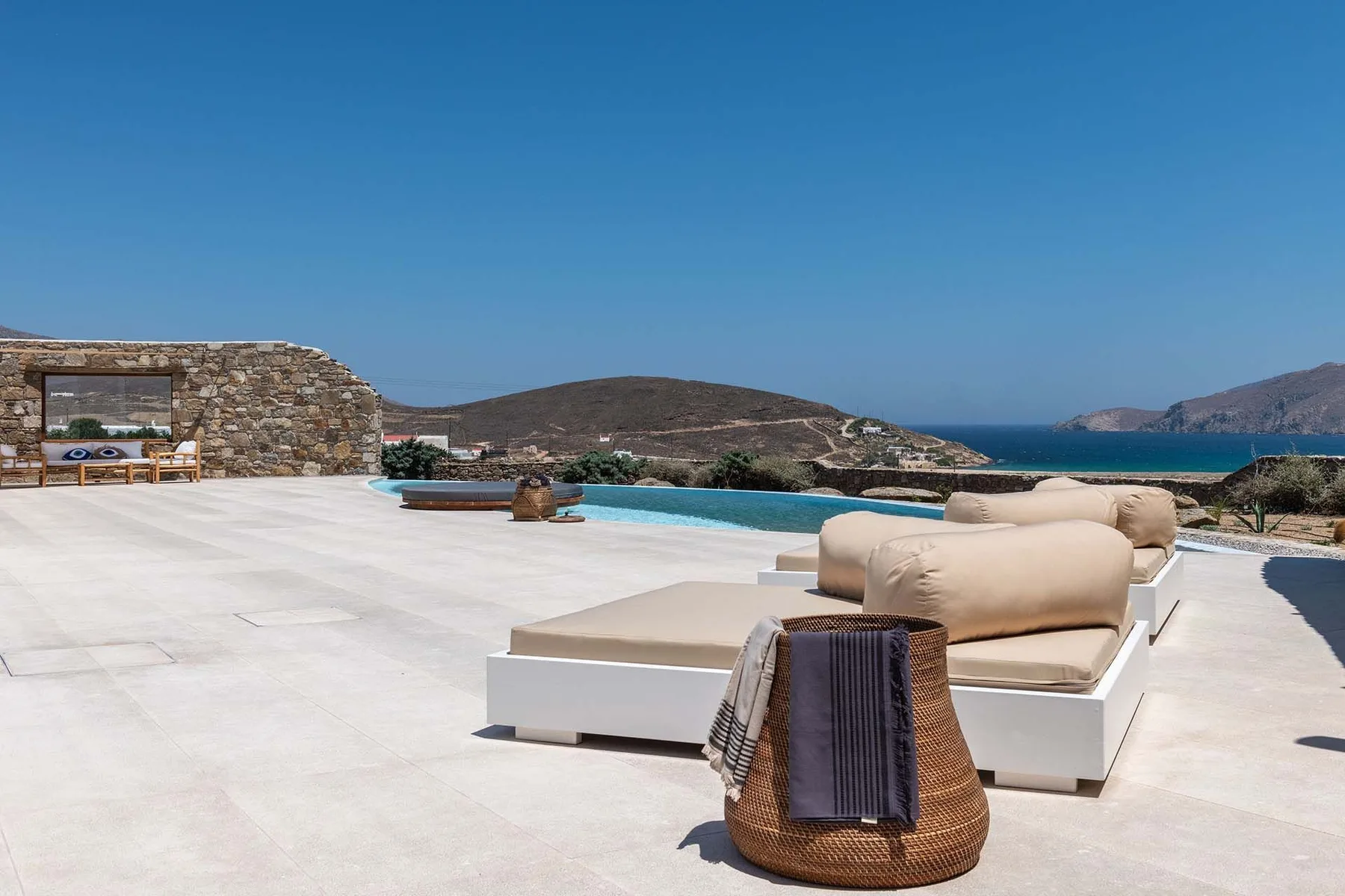 A sunlit outdoor terrace has cushioned seating beside a swimming pool, with hills and sea beyond.