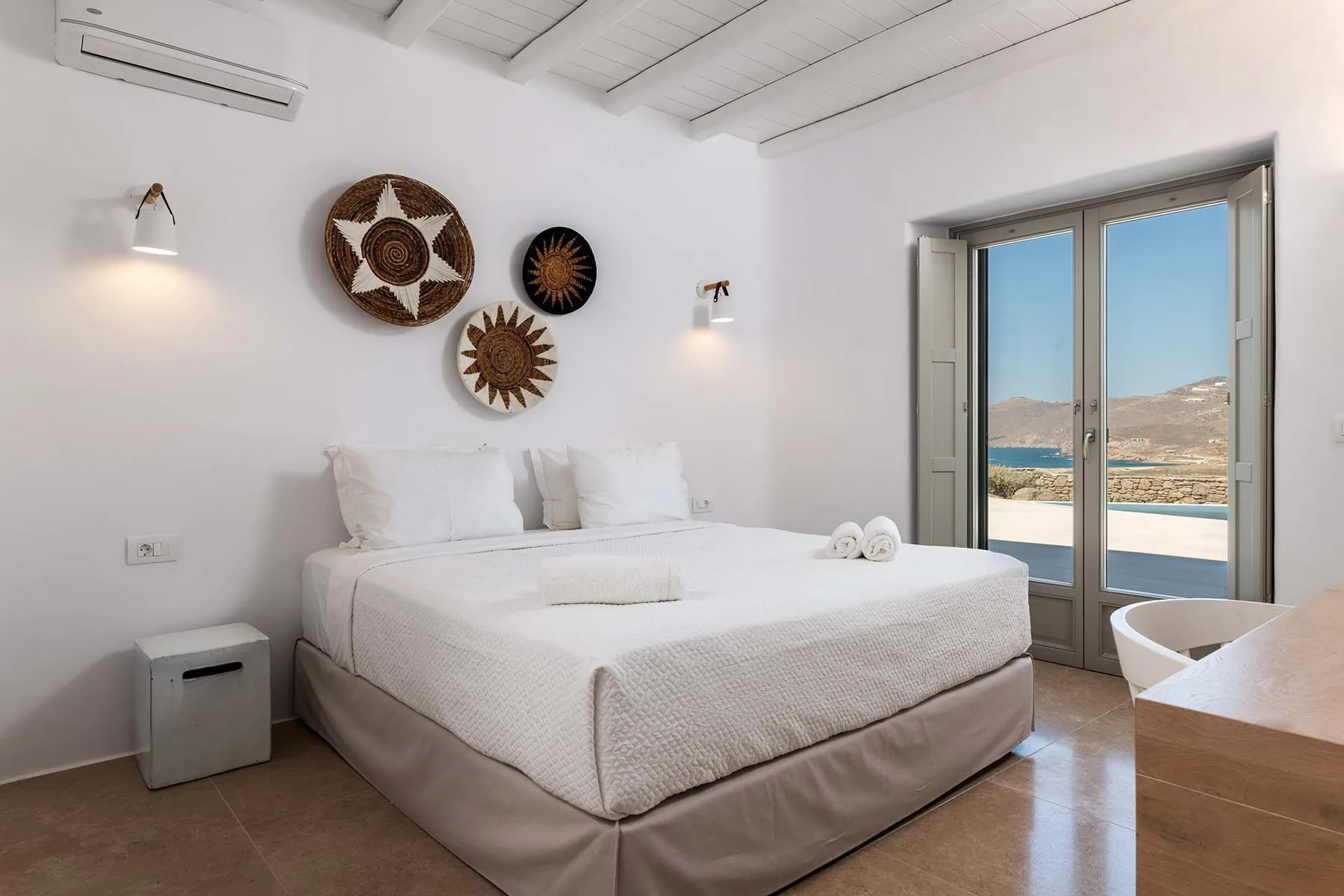 A bright bedroom with a neatly made bed and glass doors opening onto a sea view.